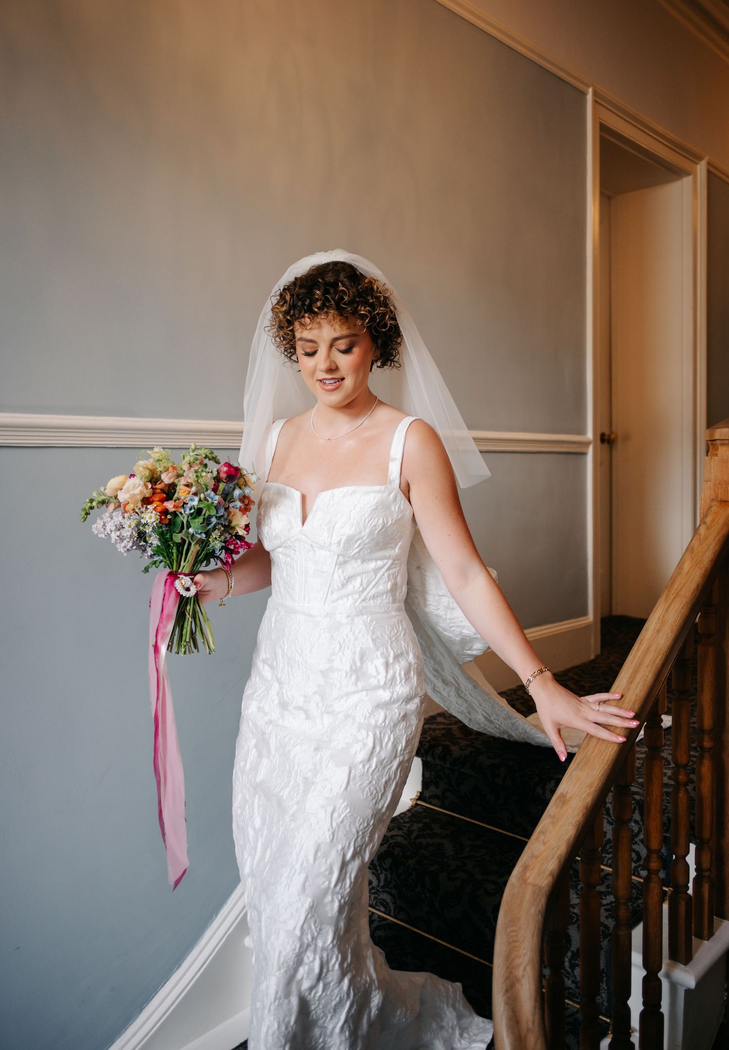 A person in a fitted lace wedding dress and veil stands on a staircase holding a floral bouquet.
