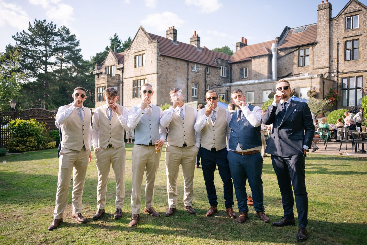 Groomsmen and wedding party members pose together in khaki suits on a green lawn outside a stone mansion.
