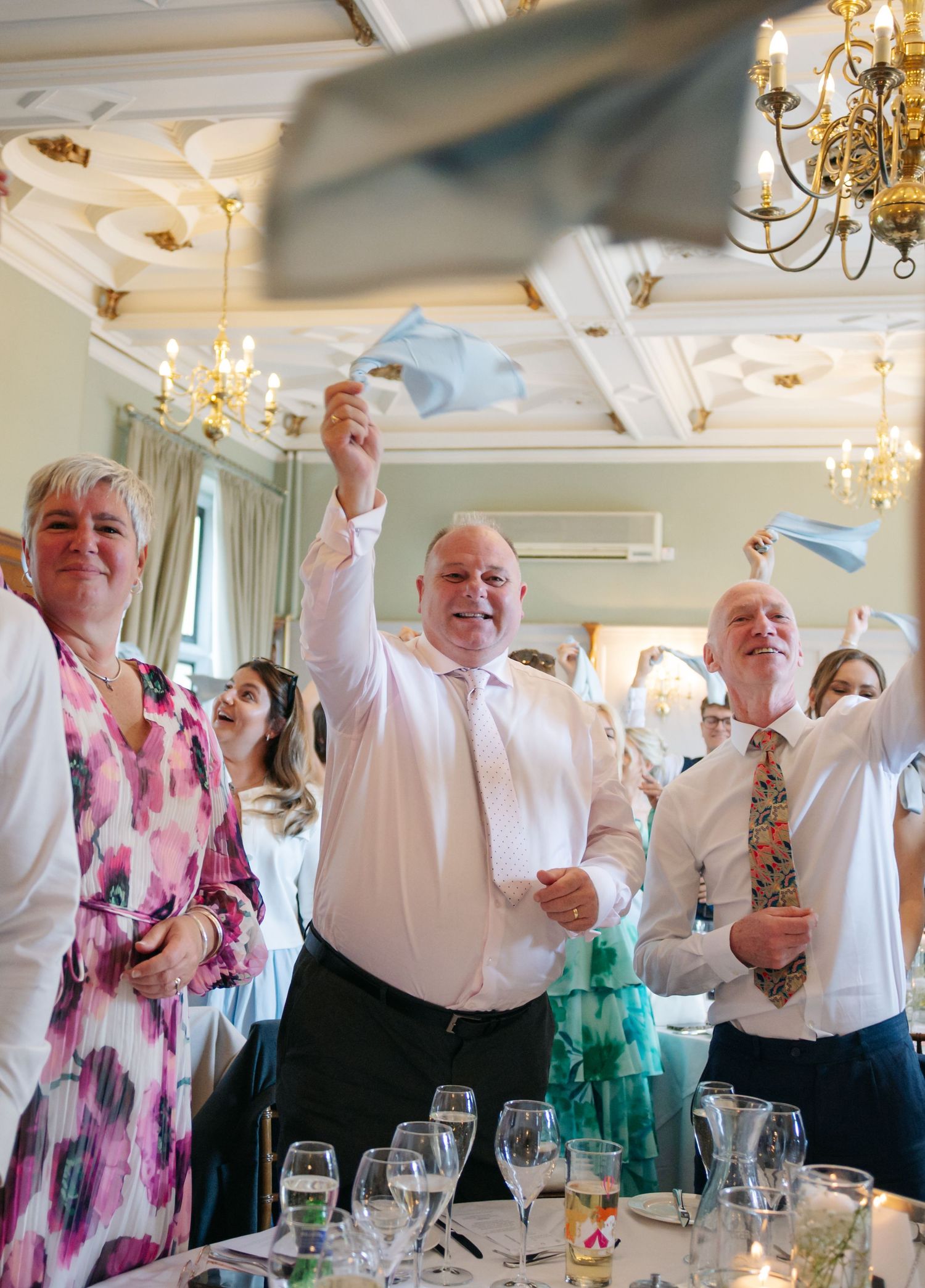 Wedding reception guests celebrate joyfully with raised arms in an elegant chandelier-lit banquet room.