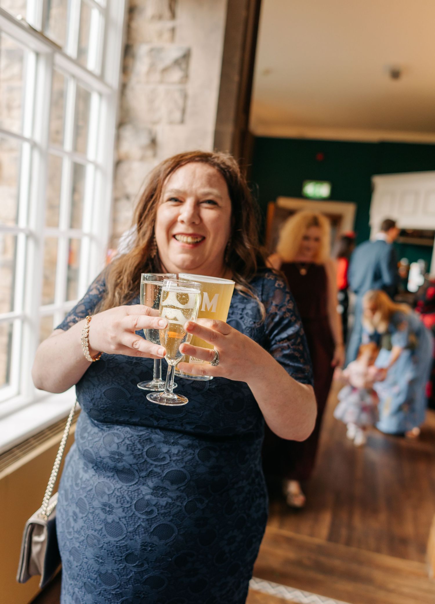 Wedding guest in navy dress making a cheerful toast with a glass of champagne by large windows.