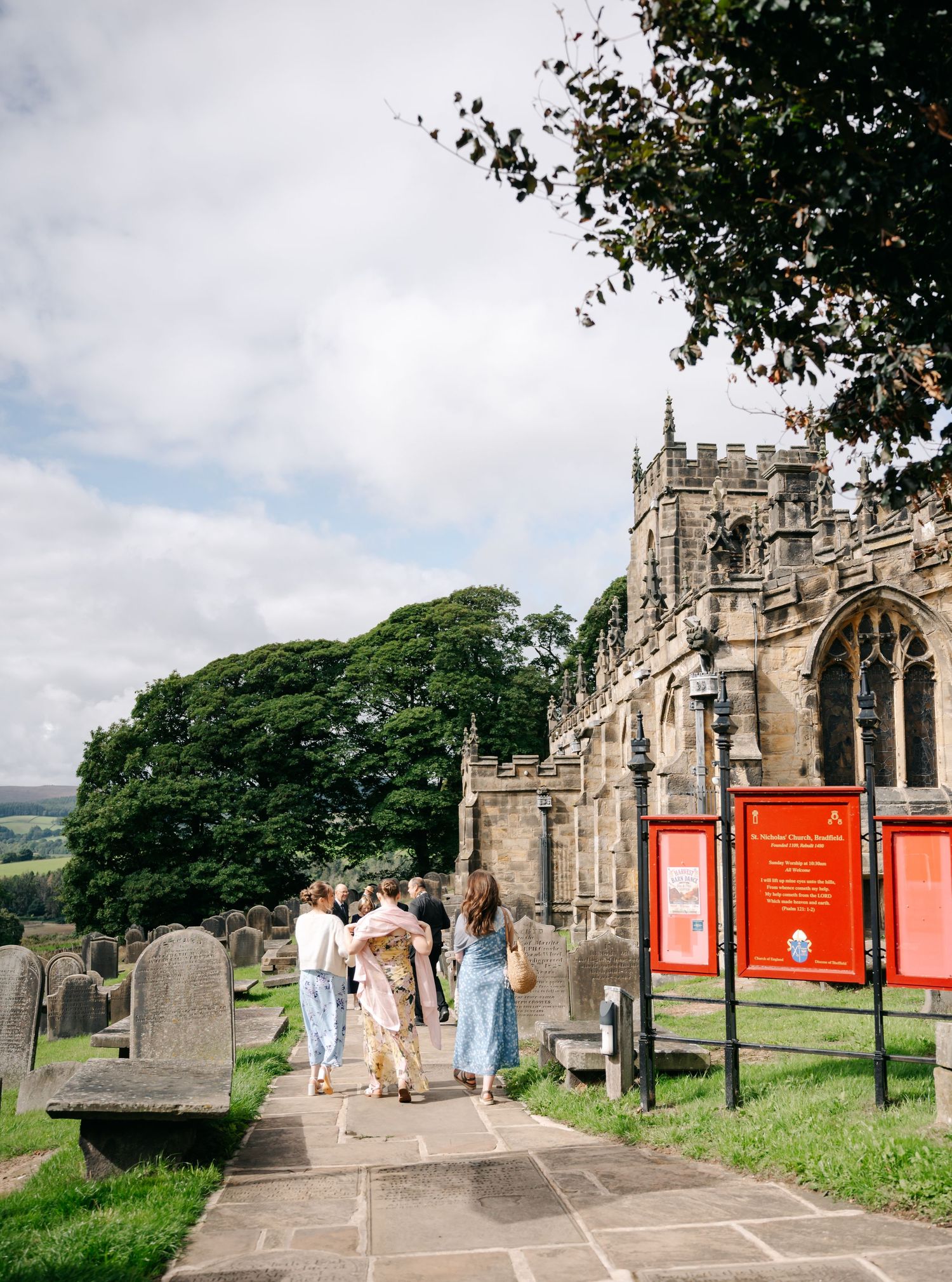Wedding guests stroll along a stone path through the churchyard toward a historic church beneath a bright cloudy sky.