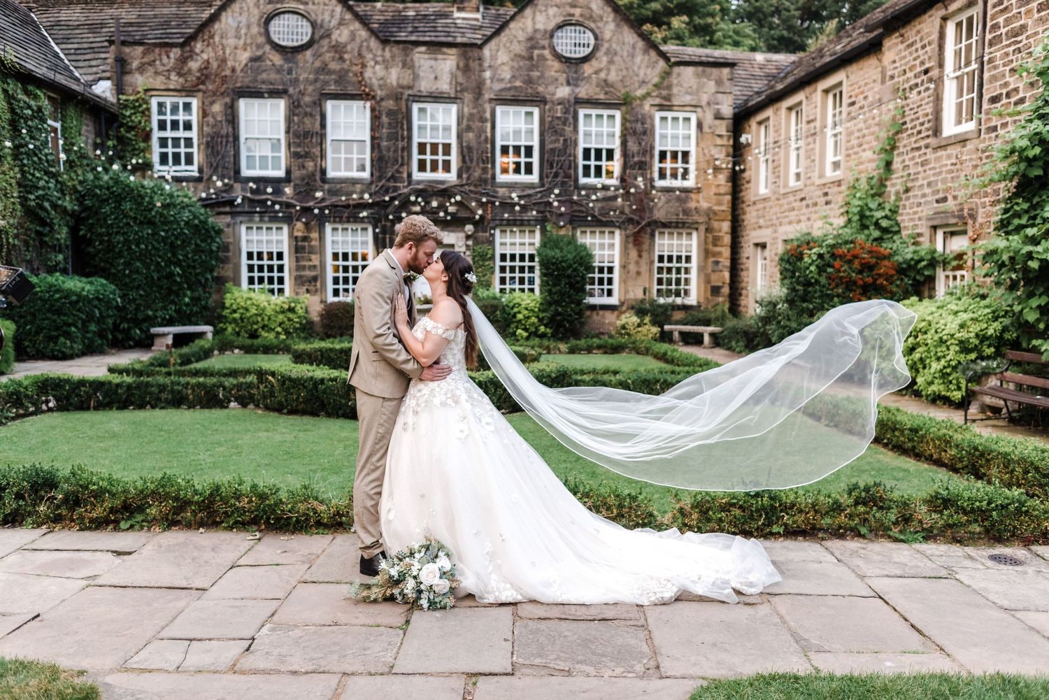 Long bridal veil flowing in wind against historic stone mansion with manicured gardens.