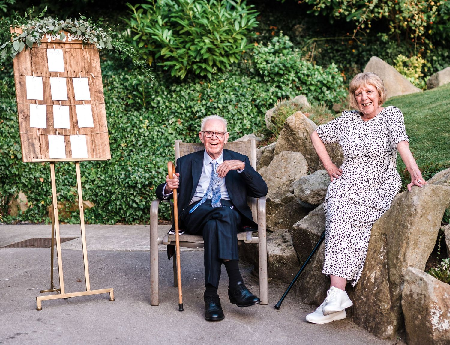 Outdoor wedding display with wooden seating arrangement board and guests relaxing on rocks.