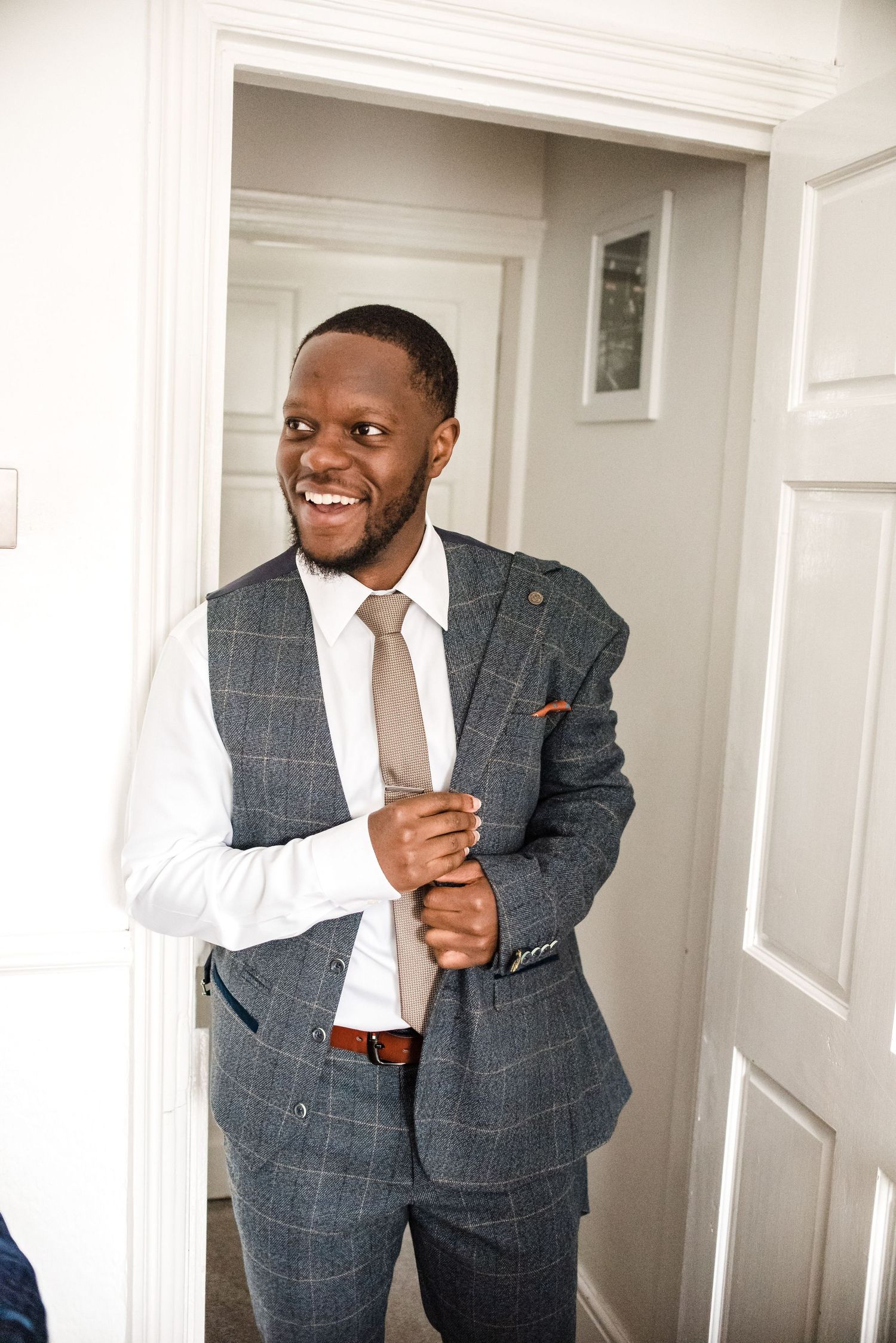 A person wearing a gray windowpane suit smiles while adjusting their tie.
