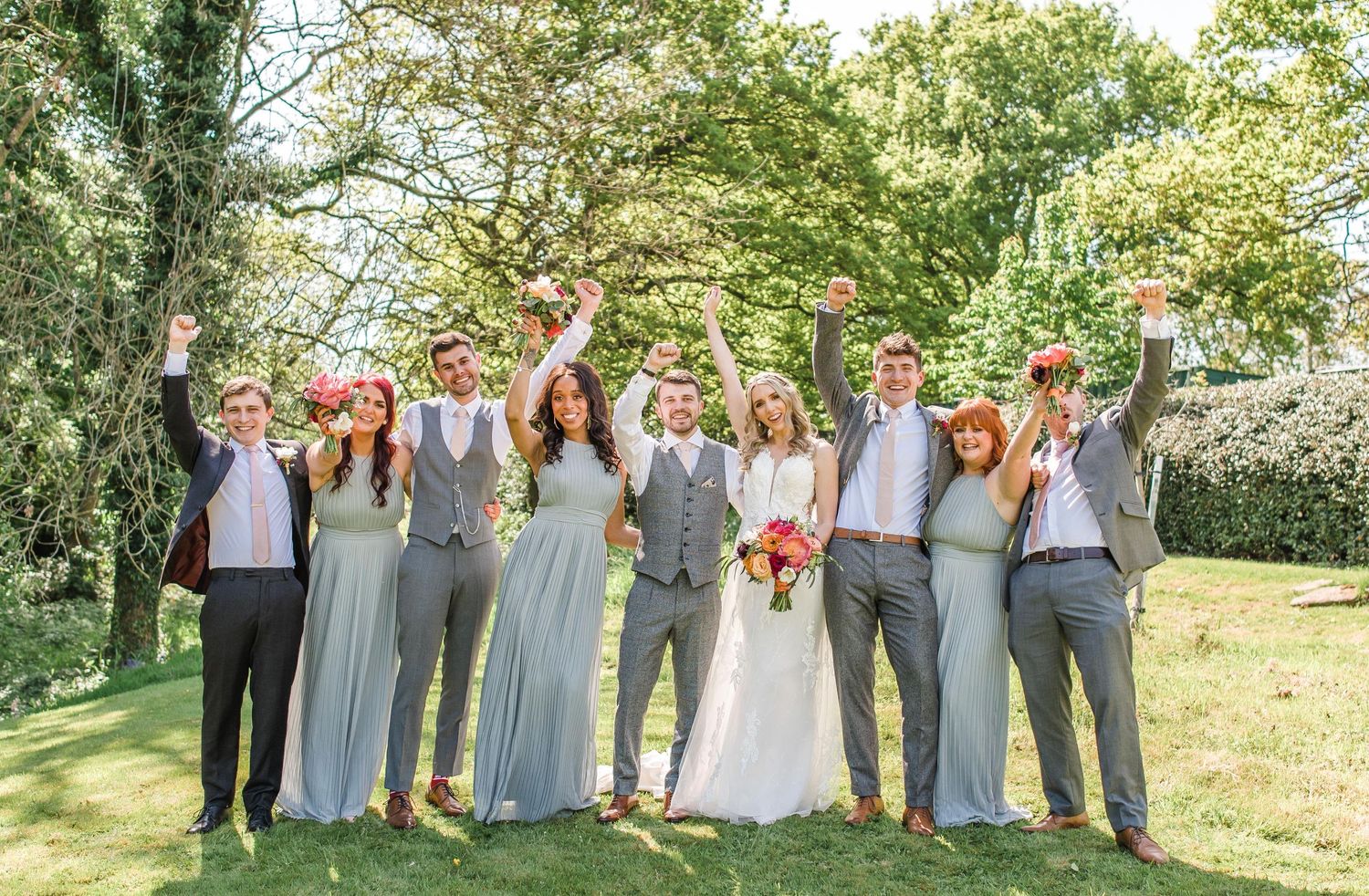 Joyful wedding party raising bouquets in celebration on sunny lawn.