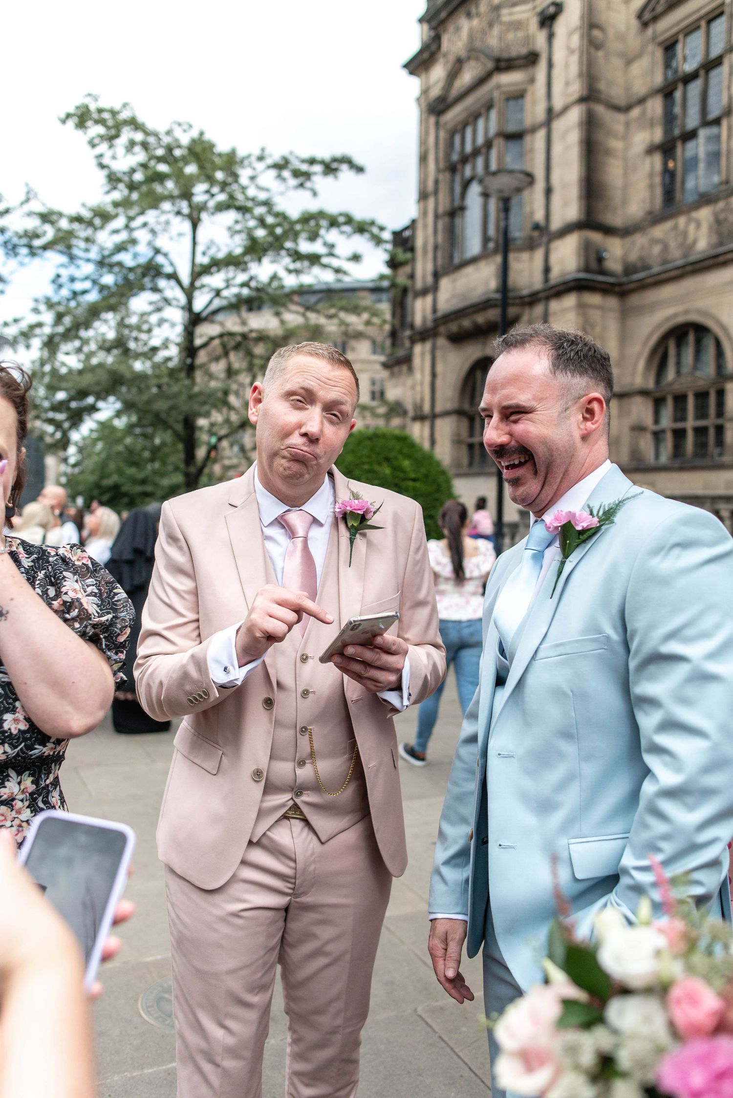 Two wedding guests in pale suits share a laugh while looking at a phone outside a historic building.