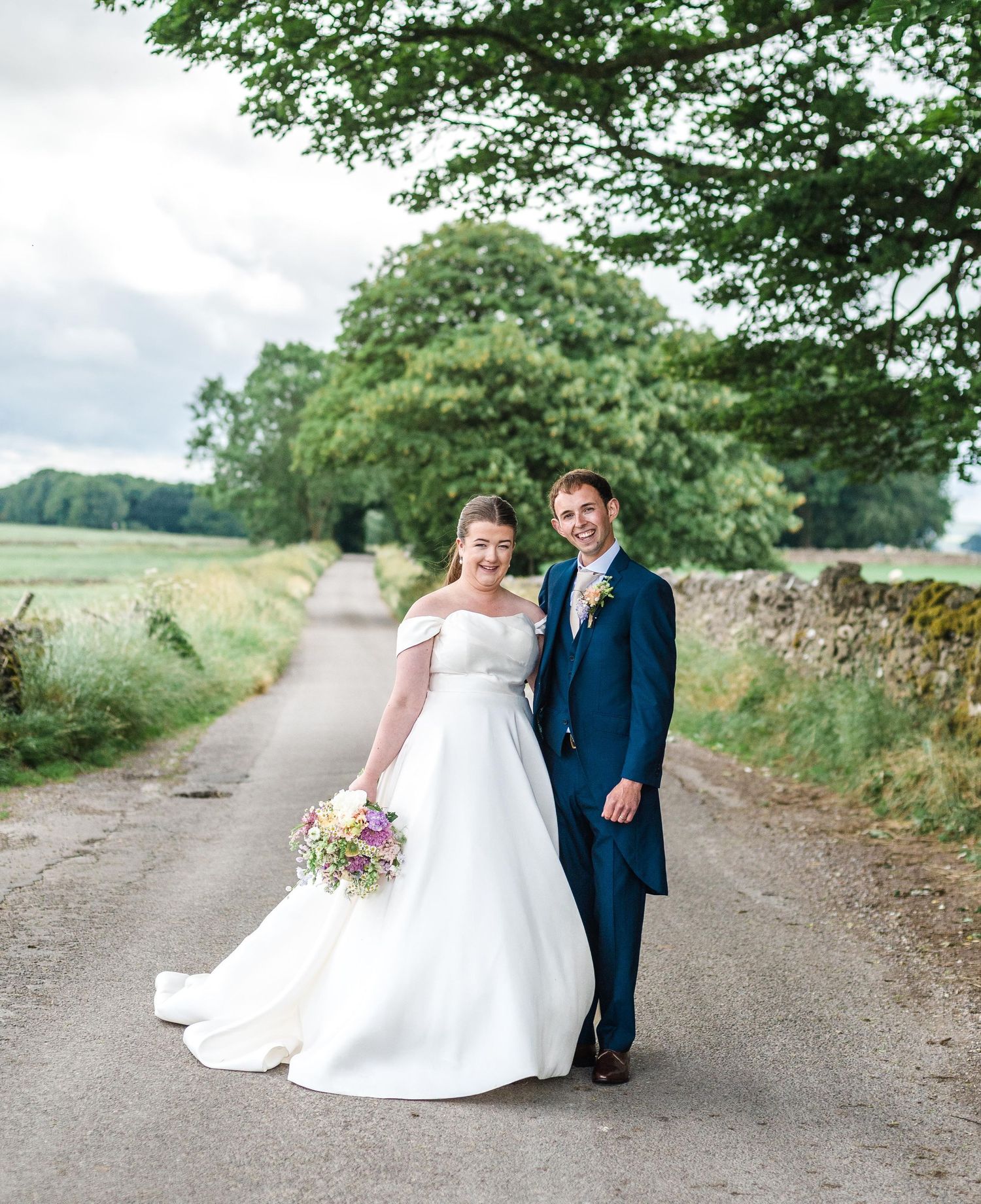 A newlywed couple poses on a country lane with trees and stone walls in the background.