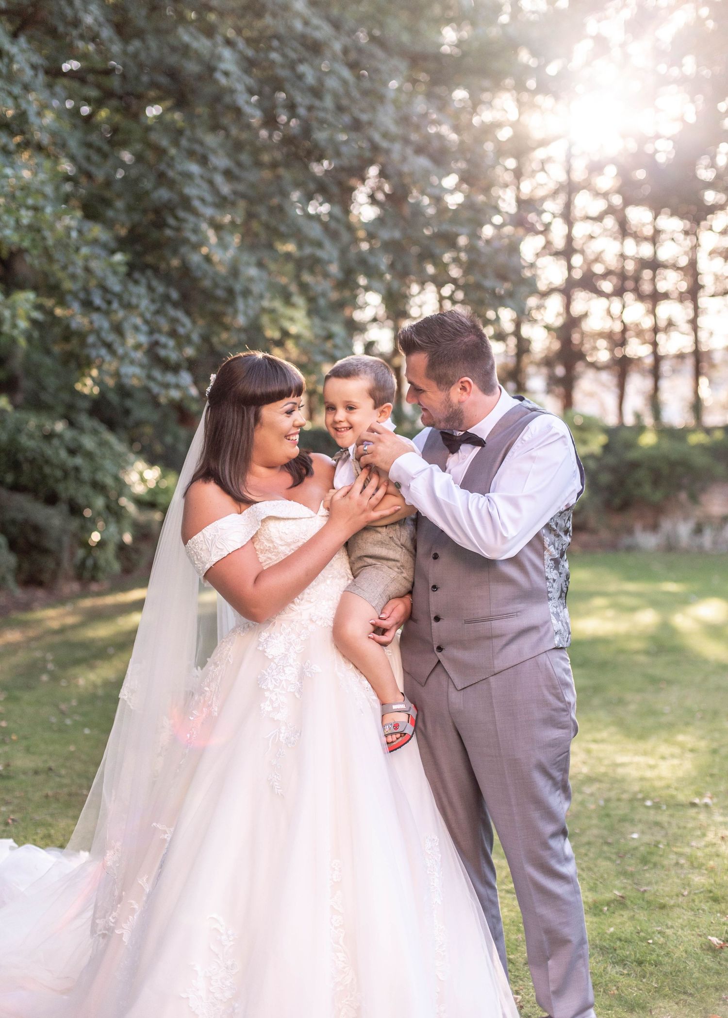 A romantic wedding moment in a sunlit garden with a couple in formal attire holding a small child.