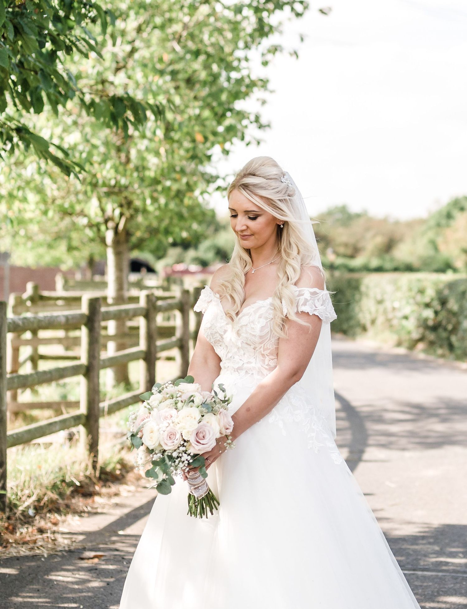Bride in off-shoulder lace wedding gown poses with bouquet on country path.