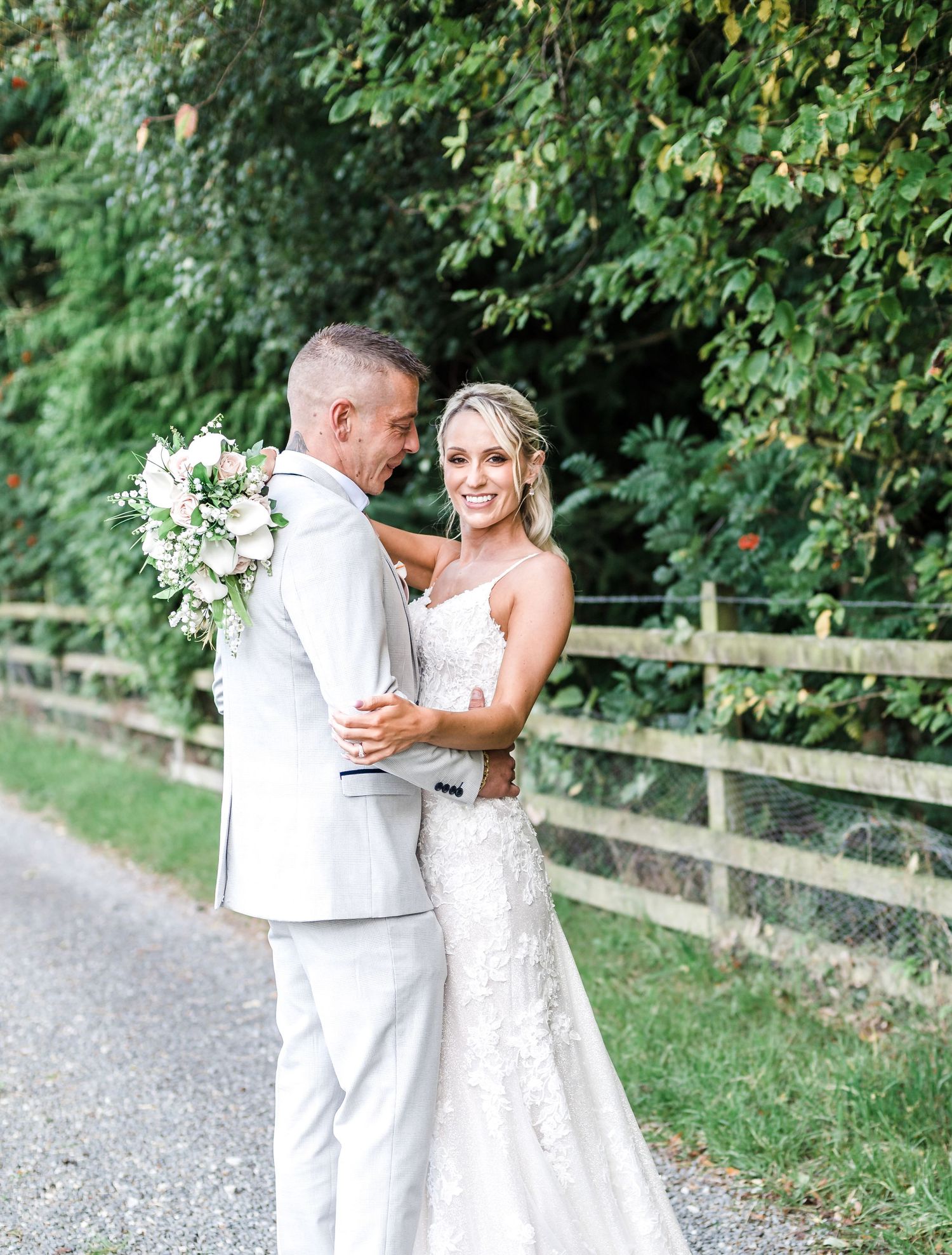 Elegant wedding couple poses on a country path surrounded by green foliage.