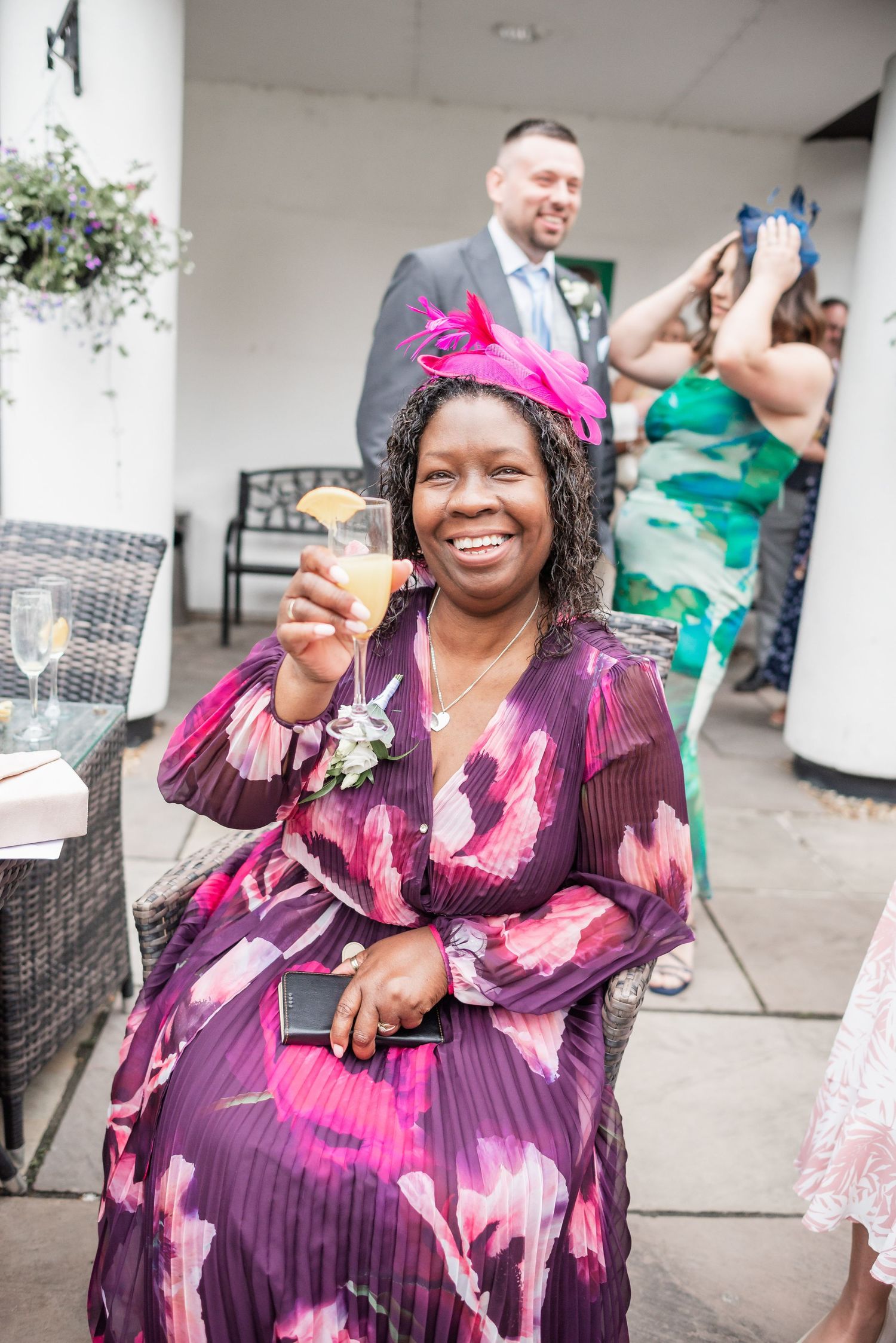 Guest in a purple floral dress and pink fascinator enjoys a drink at an outdoor wedding reception.