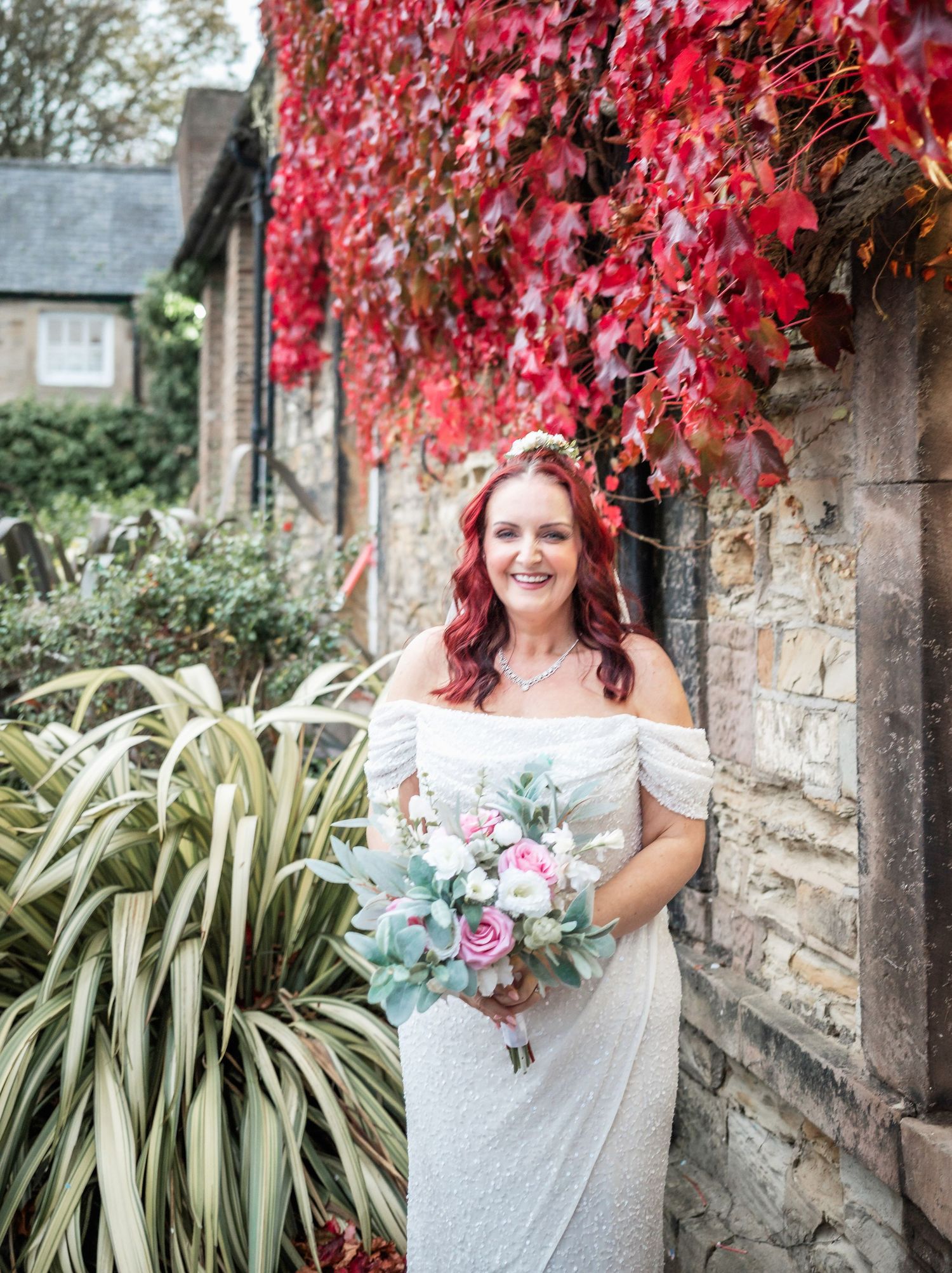 Bride poses against wall covered in vibrant red autumn leaves while holding floral bouquet.
