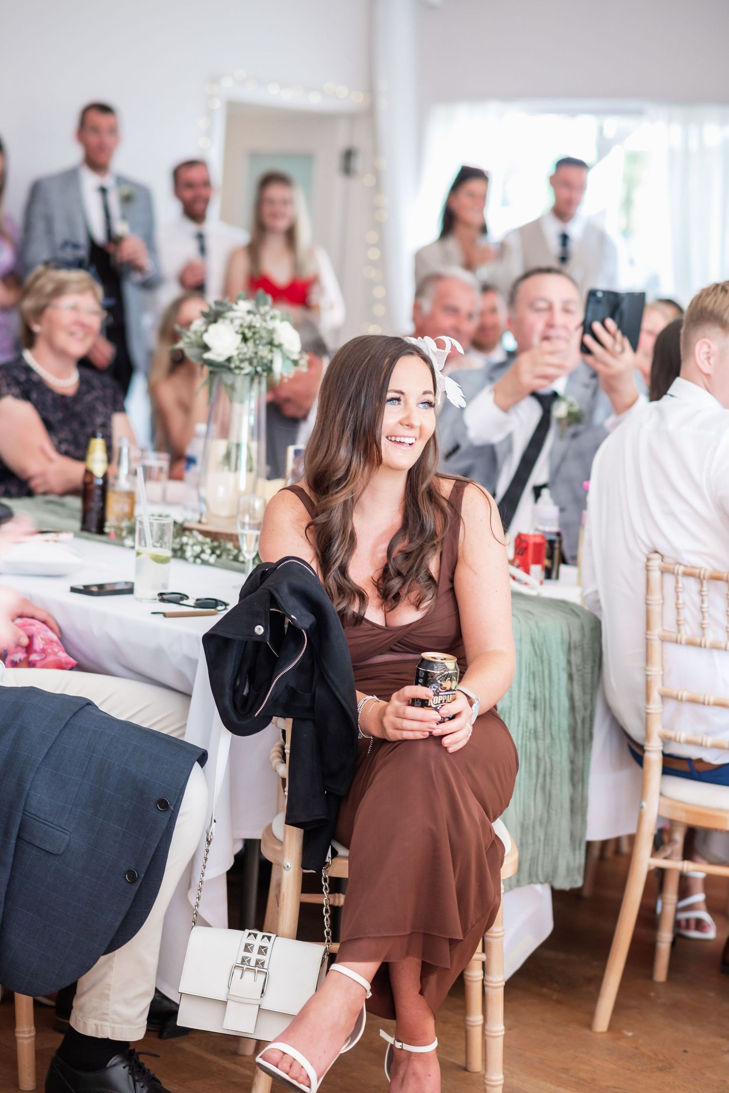 Wedding reception guest in brown dress seated at decorated table during indoor celebration.