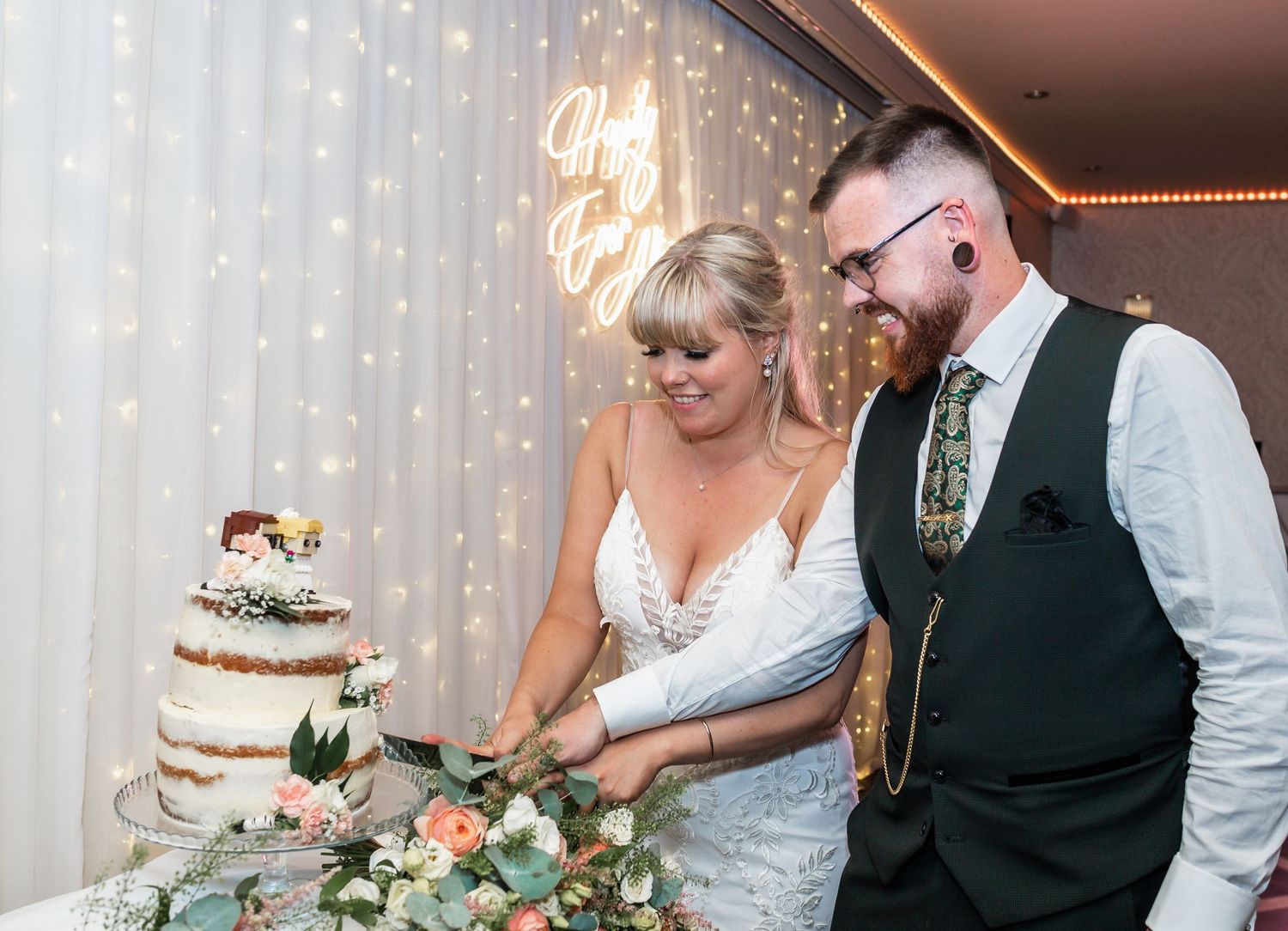 Couple cutting their elegant semi-naked wedding cake decorated with fresh greenery and flowers.