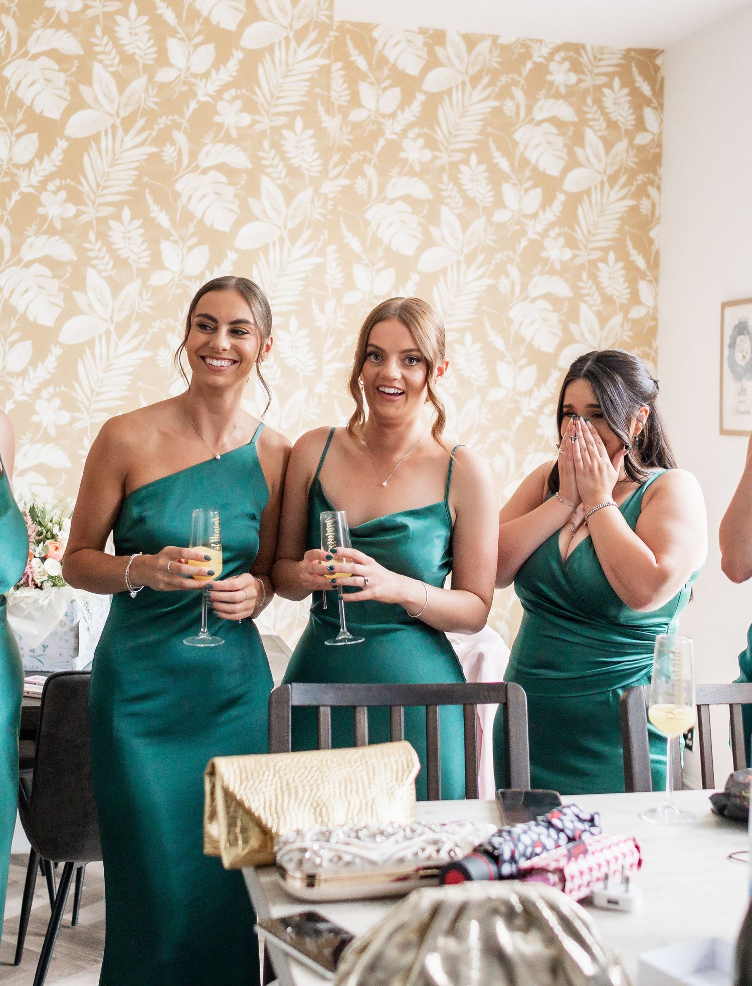 Three bridesmaids in elegant emerald green dresses share a celebratory moment with champagne glasses.