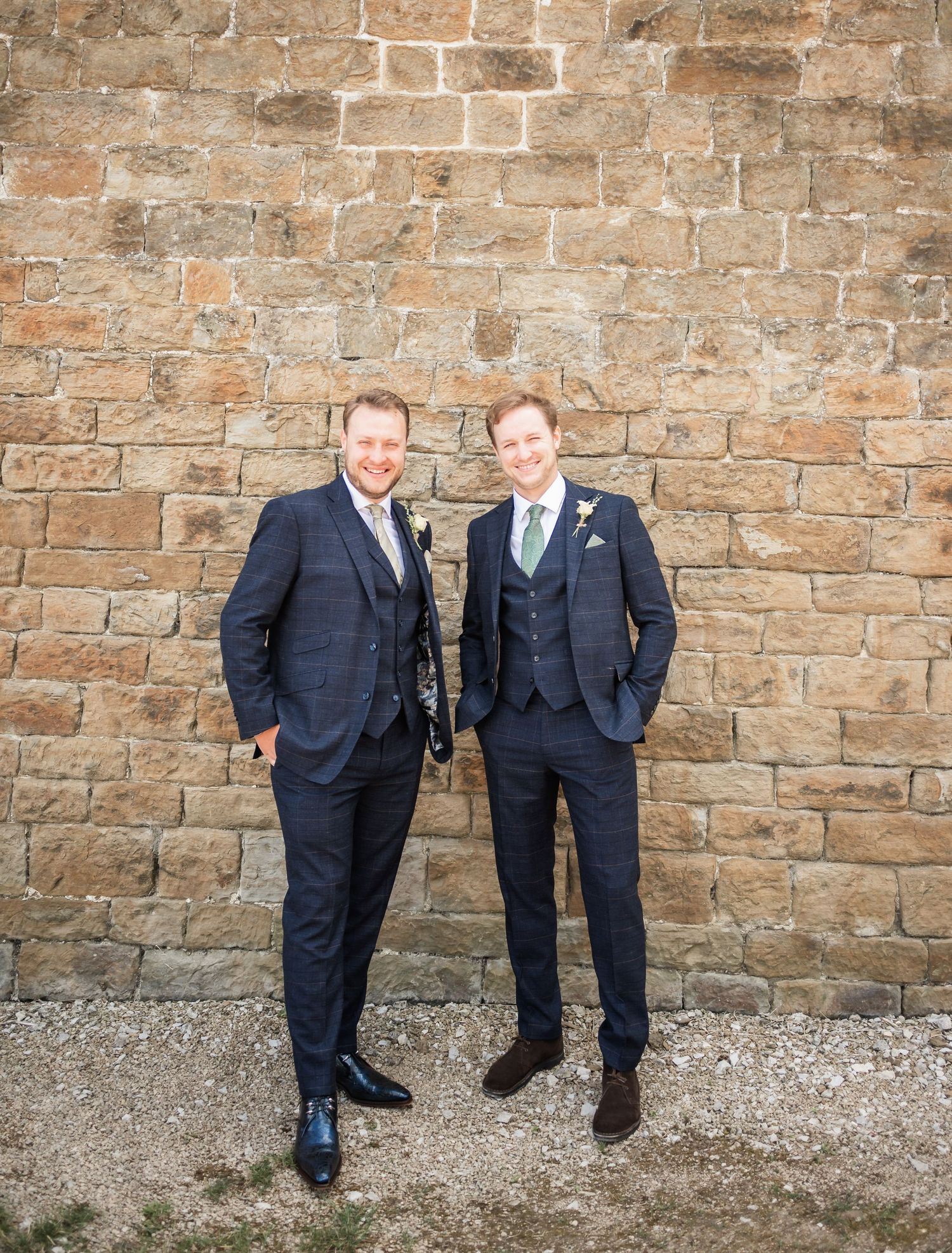 Two men in formal suits share a lighthearted moment against a stone wall background.
