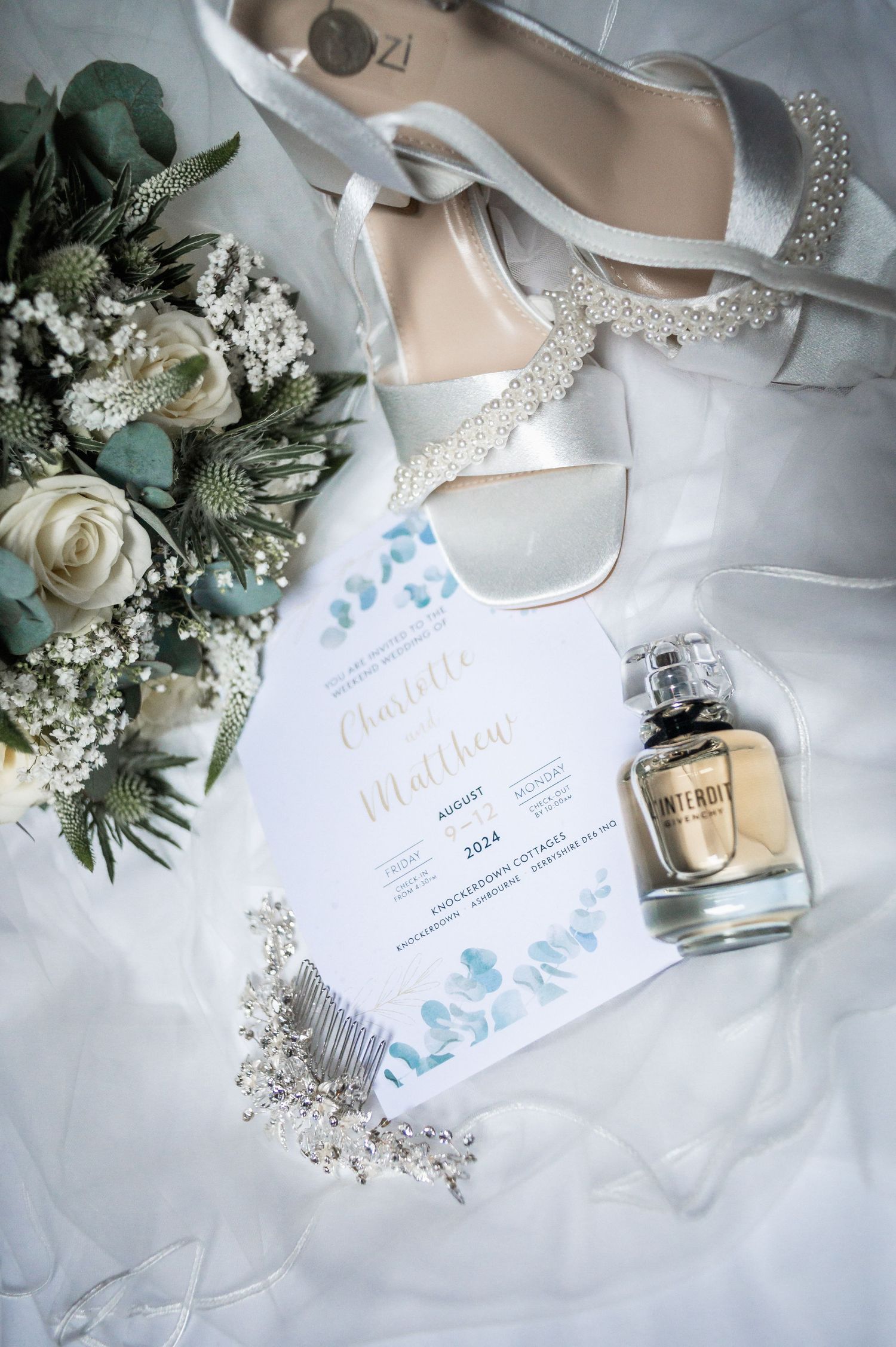 Wedding details flatlay with invitation, bridal shoes, perfume bottle and white floral bouquet.