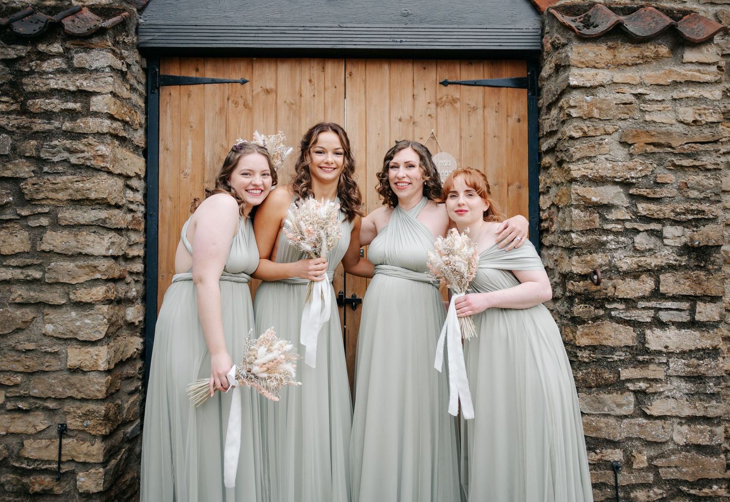 Row of bridesmaids in sage green dresses standing against a rustic stone wall.