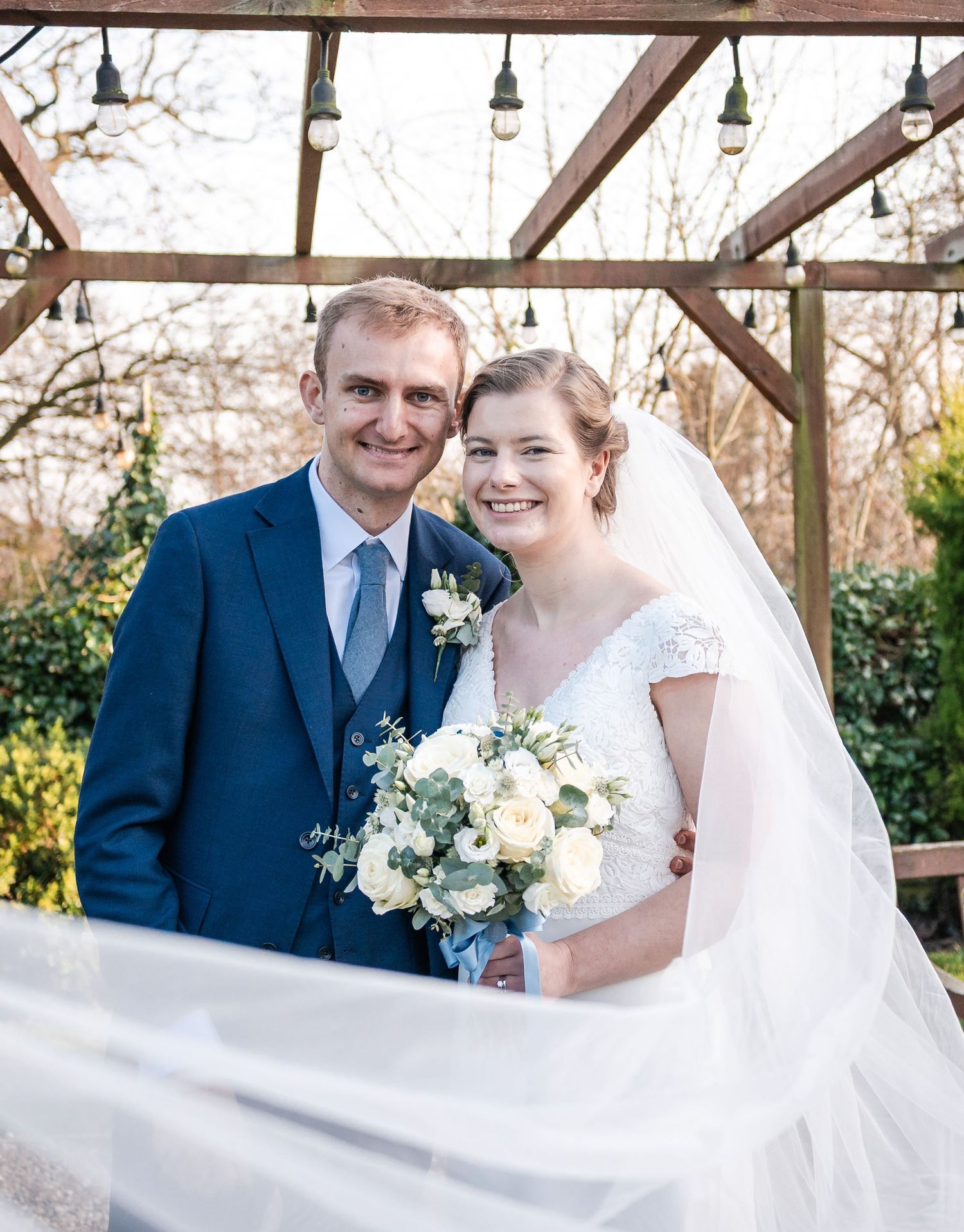 Newlyweds share romantic moments under a wooden pergola with hanging lights during their wedding celebration.