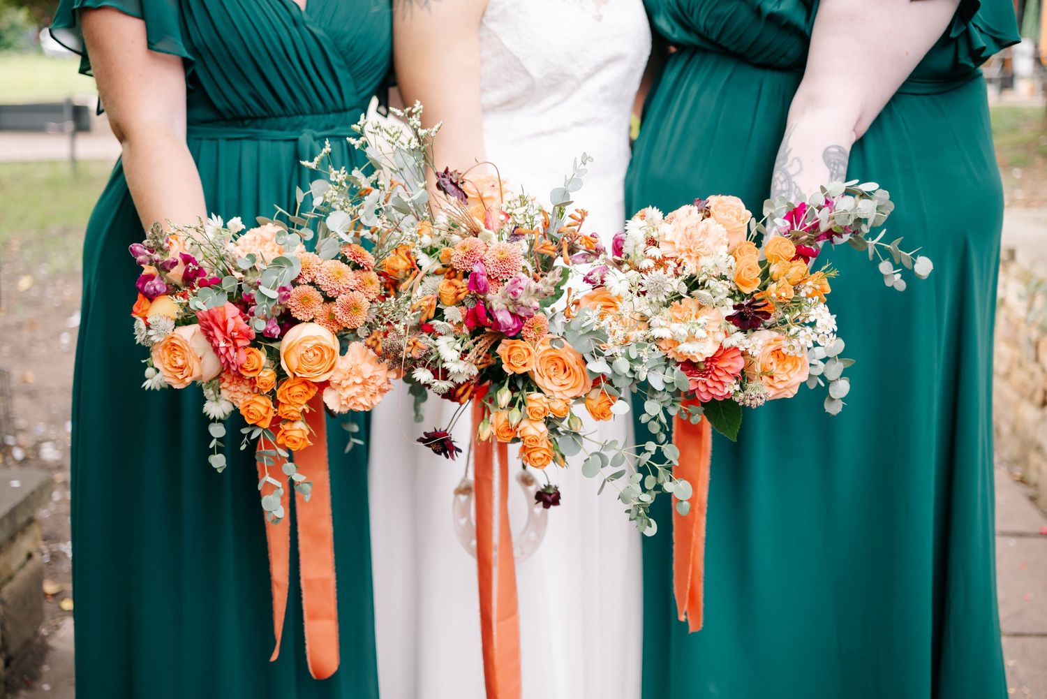 Row of emerald green bridesmaid dresses with matching orange and peach bouquets.