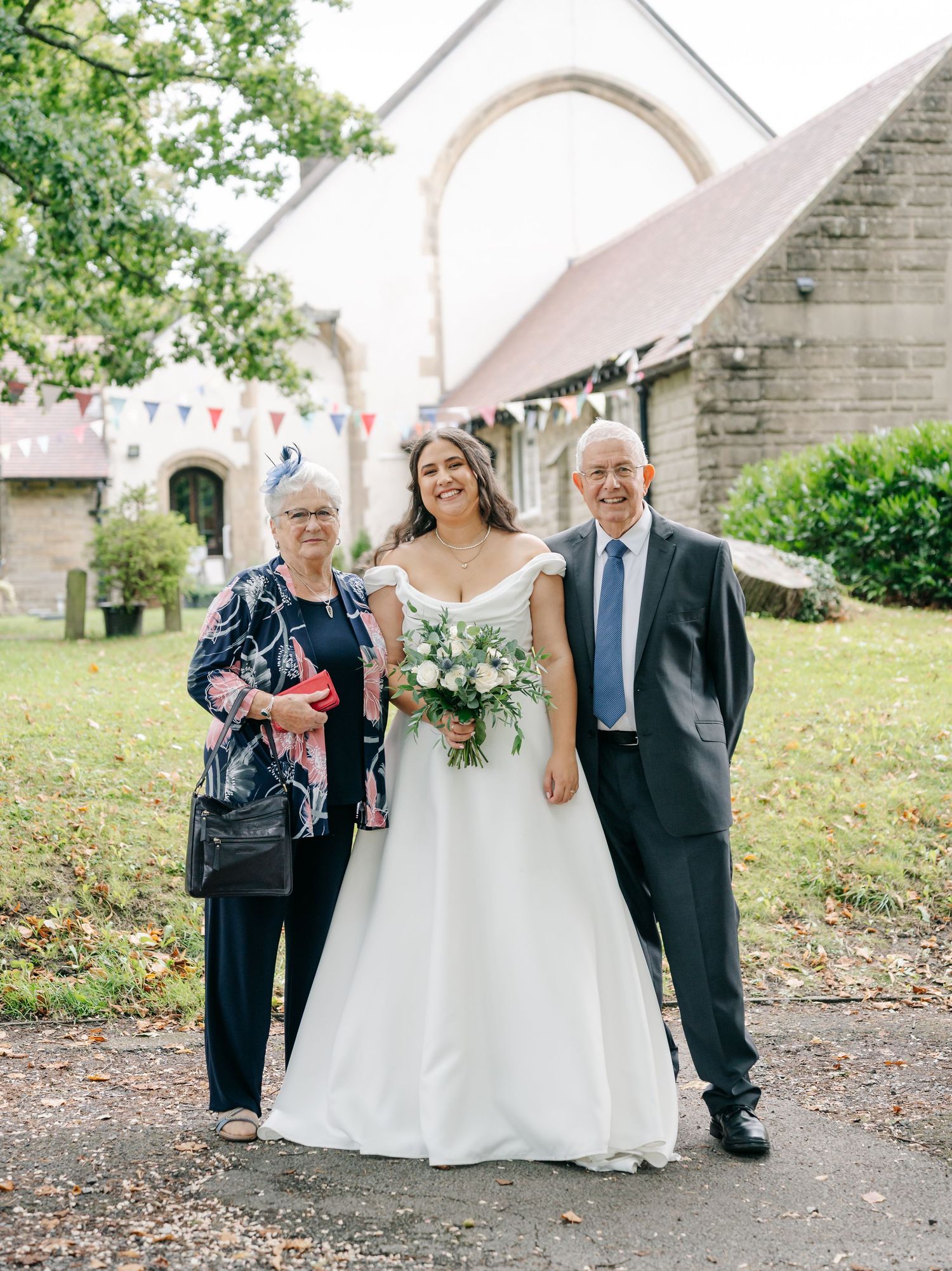 Wedding party gathered outside a stone church with white bunting decorations.