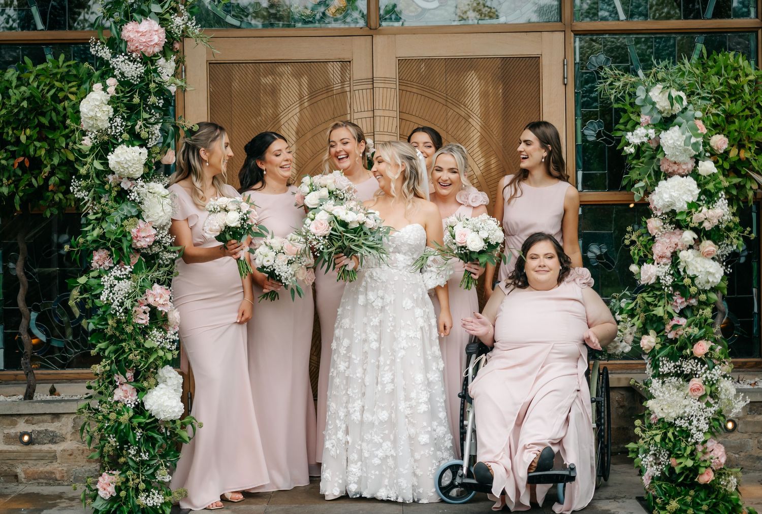 Wedding party in blush pink dresses poses outdoors with white floral arrangements and greenery archway decorations.