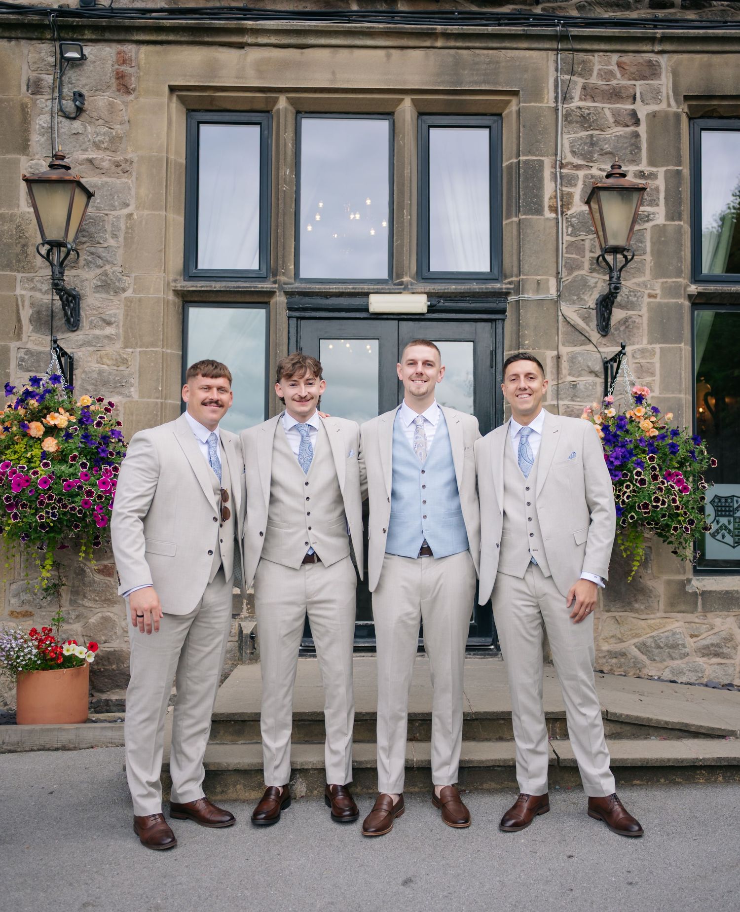 Groomsmen in light gray suits and blue ties pose together outside a stone building with flower baskets.