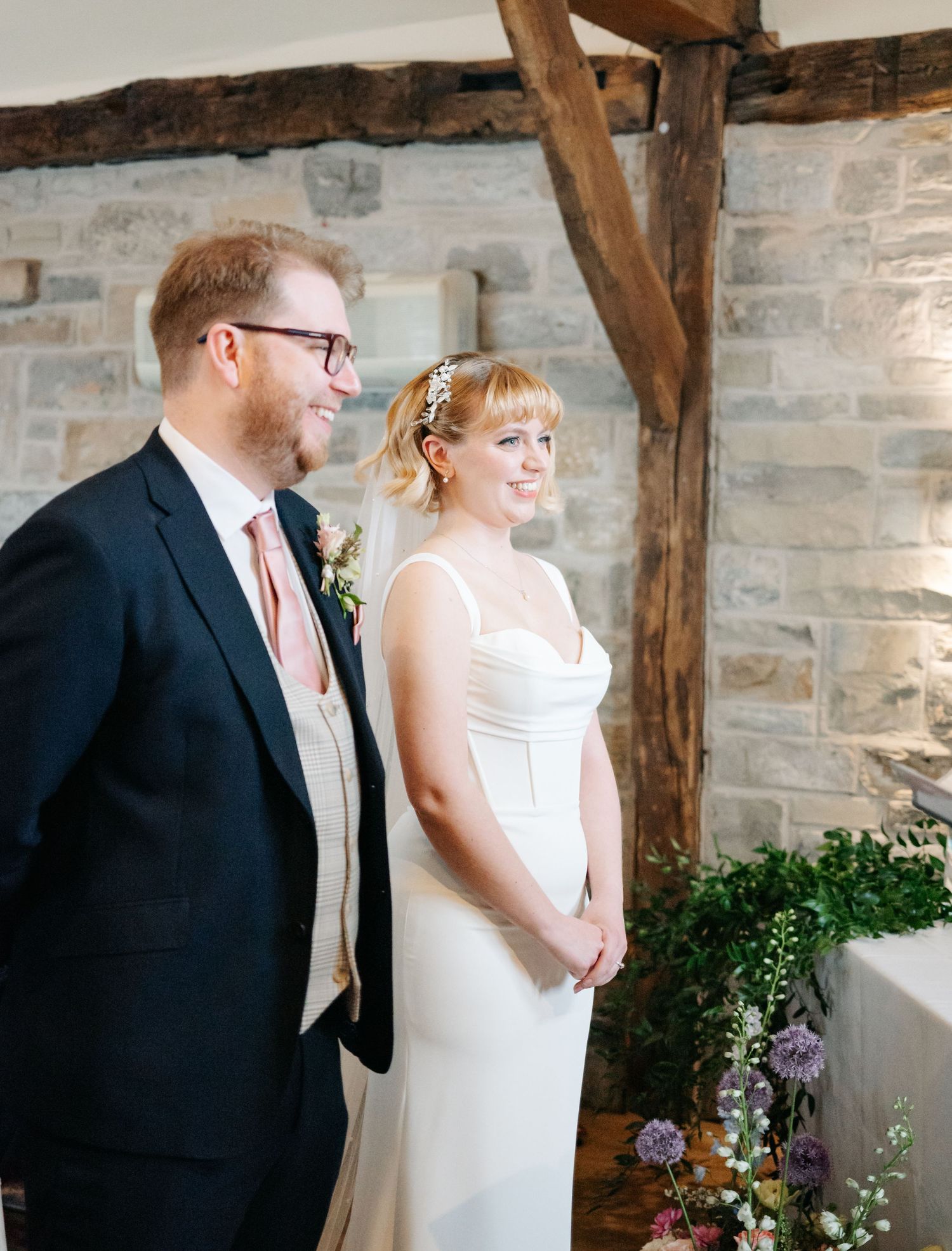 Bride and groom smiling during wedding ceremony in rustic stone venue with wooden beams and floral decorations.
