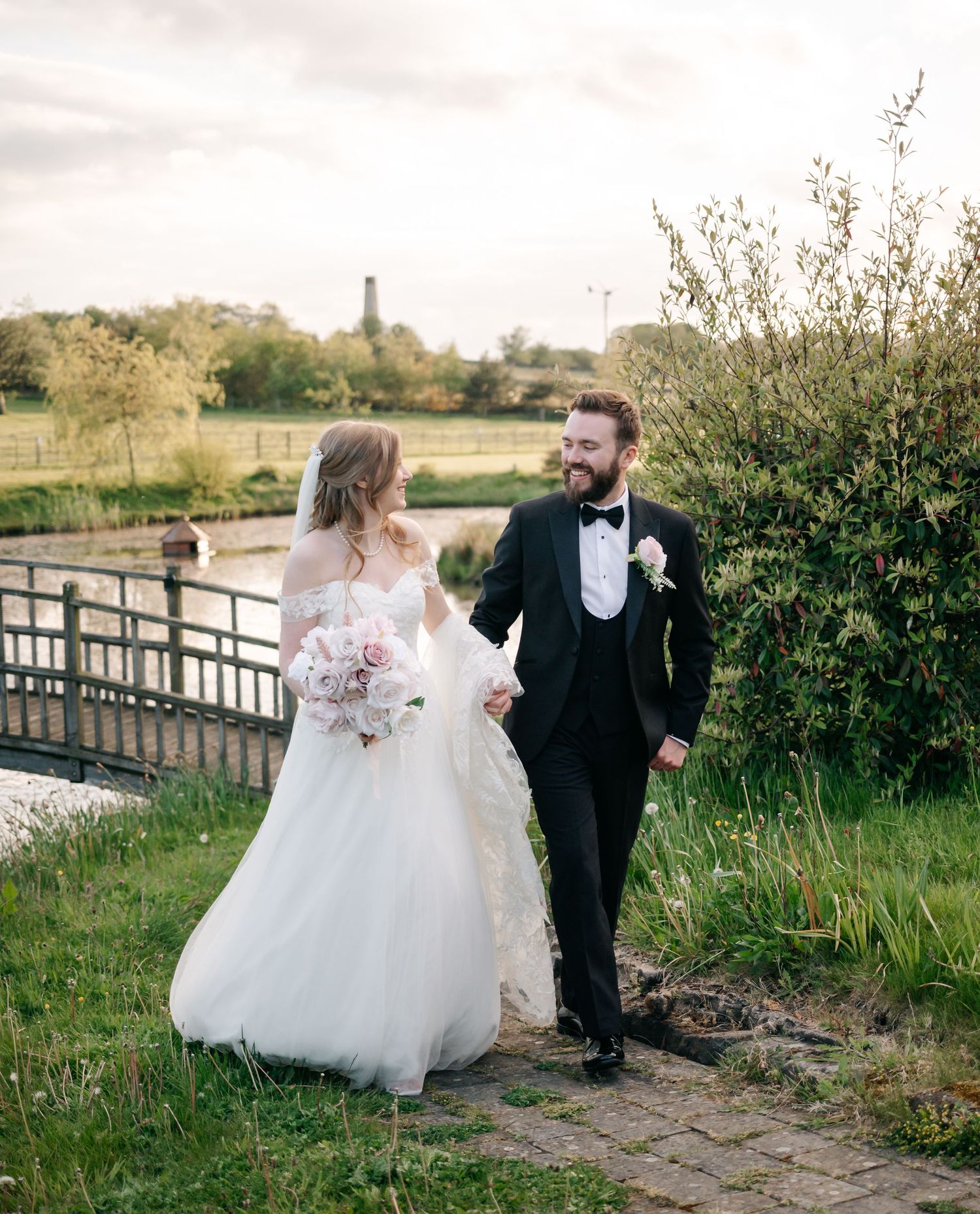 A bride and groom walk hand in hand along a garden path near a river, dressed in a white gown and black tuxedo.