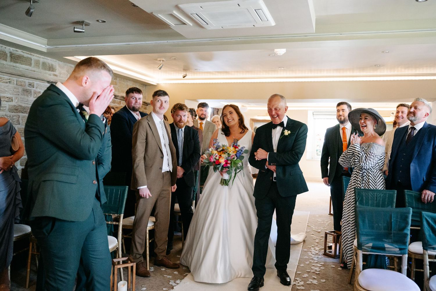 Emotional groom reacts as bride walks down the aisle with her father during an indoor wedding ceremony.