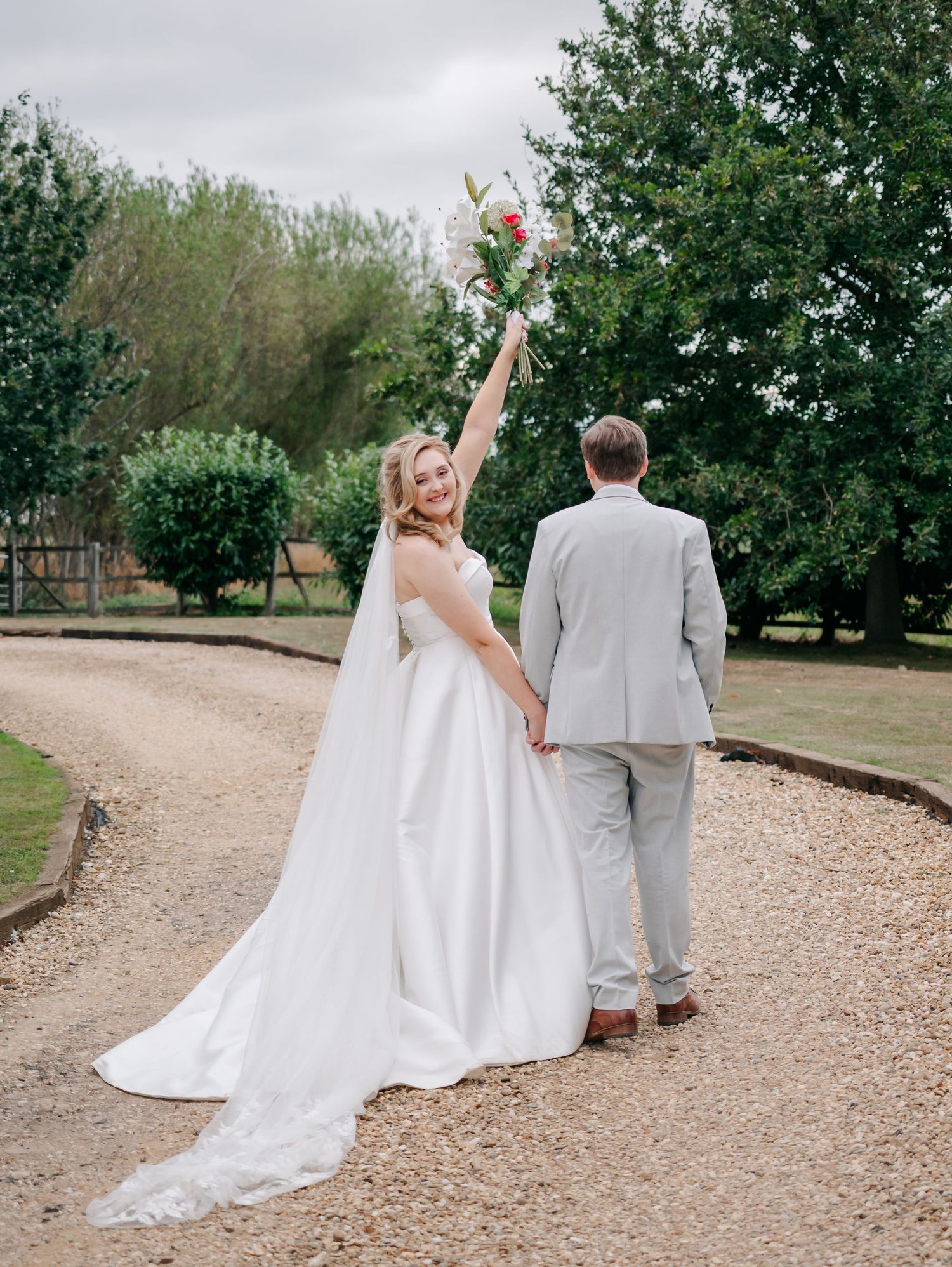 Newlyweds walking hand in hand down a gravel path surrounded by lush greenery.