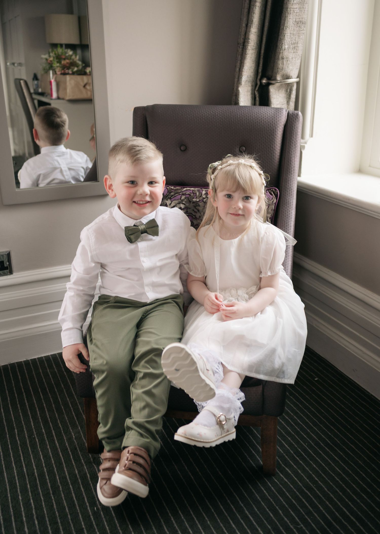 Two children in formal attire sit together on a grey armchair during a wedding celebration.