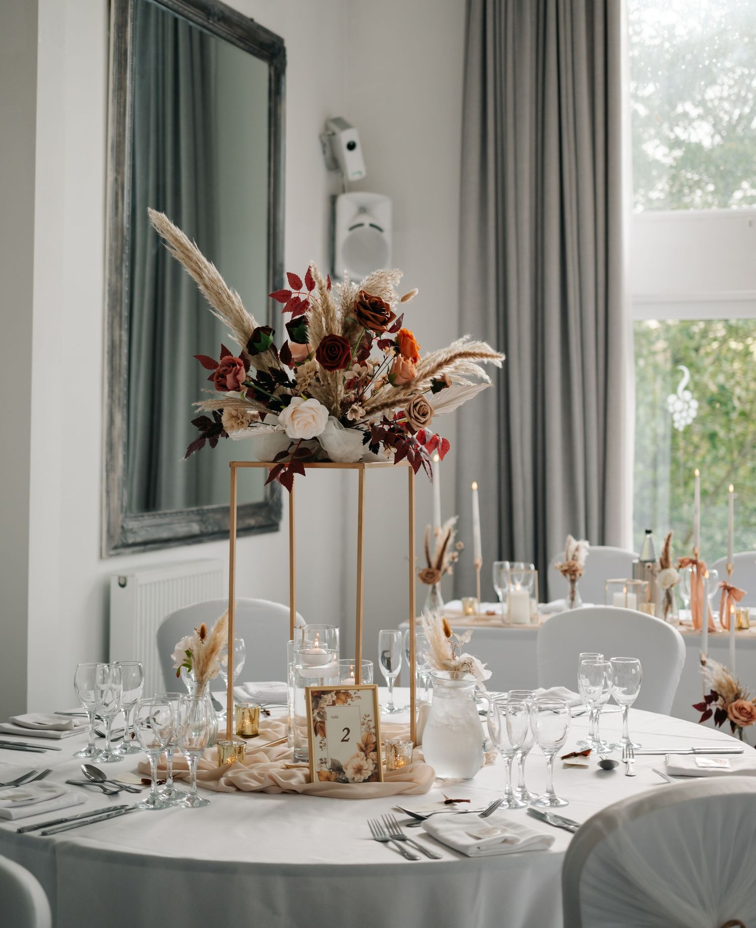 Modern wedding reception tables with tall floral centerpieces and elegant place settings in a bright room.