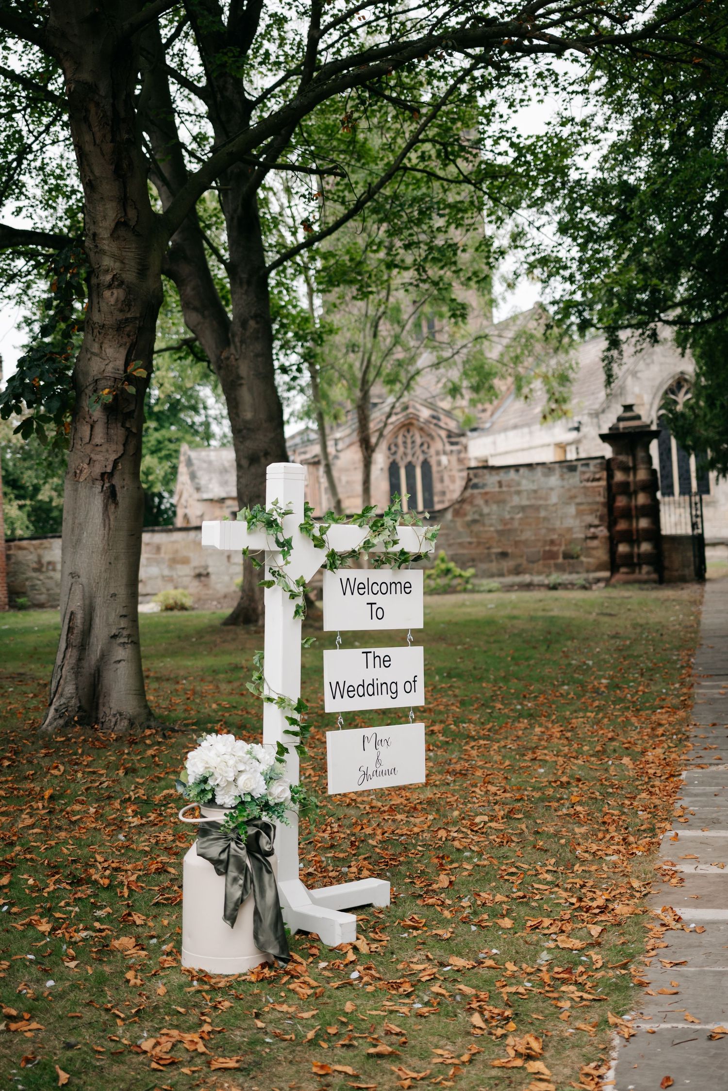 Rustic wedding welcome signs with white flowers line a tree-covered path with scattered autumn leaves.