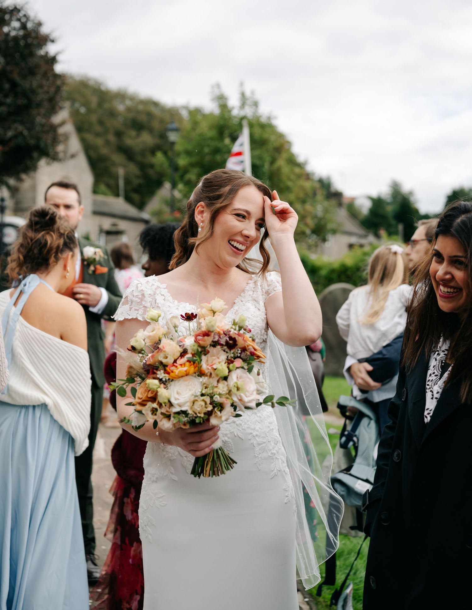 Smiling bride in a white lace gown and veil holds a colorful bouquet while guests laugh and gather outdoors at a countryside wedding.