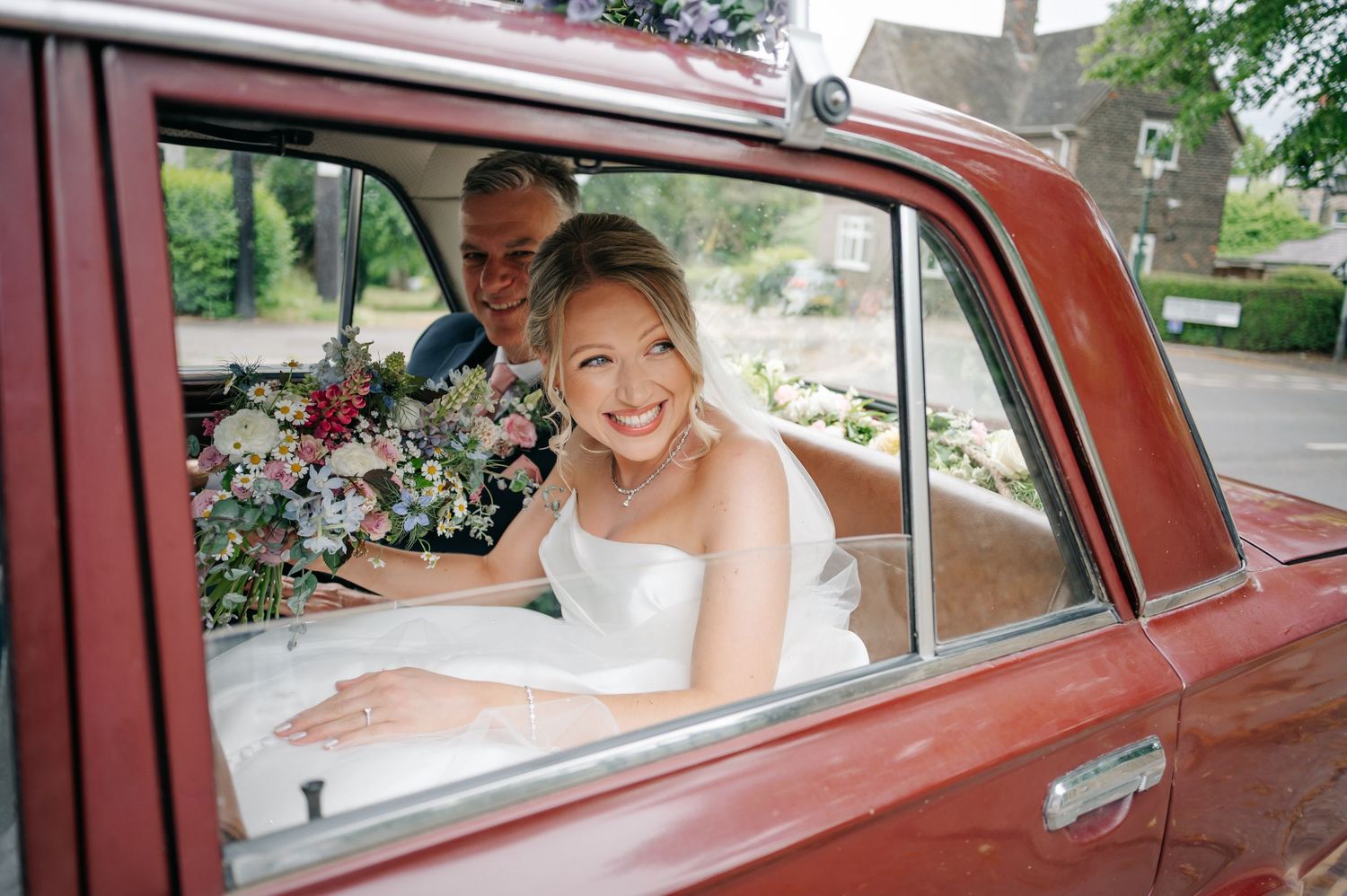 A person sits in a vintage red car smiling through the window during a wedding celebration.