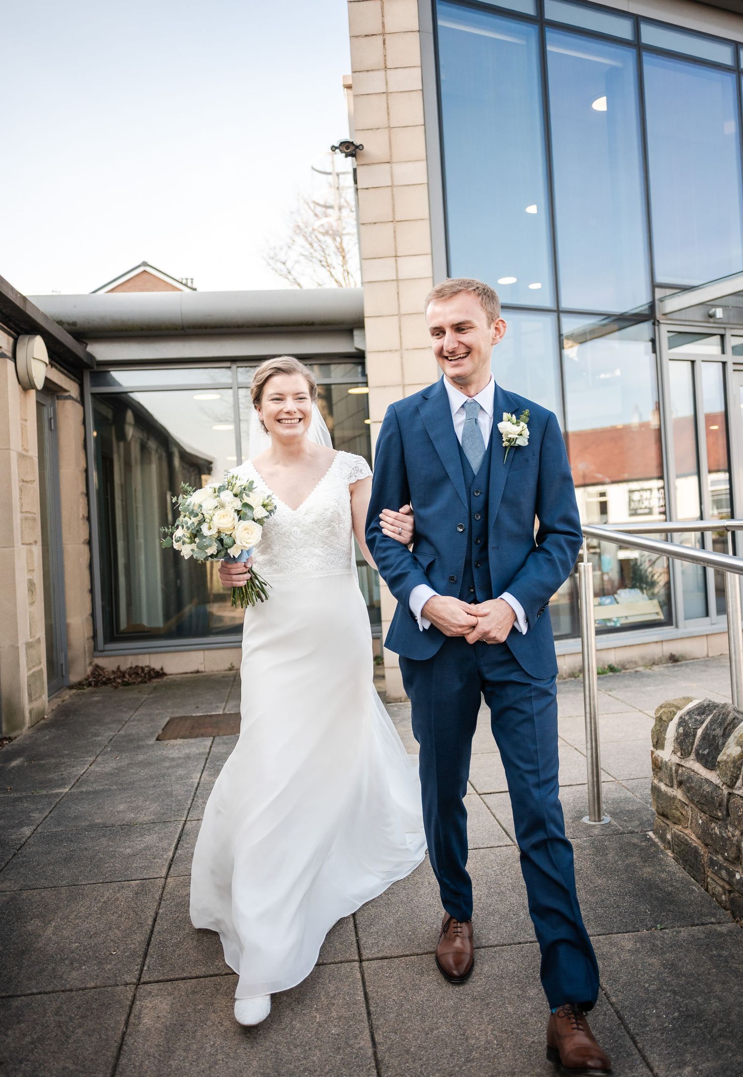 A newlywed couple walking together outside a modern building on their wedding day.