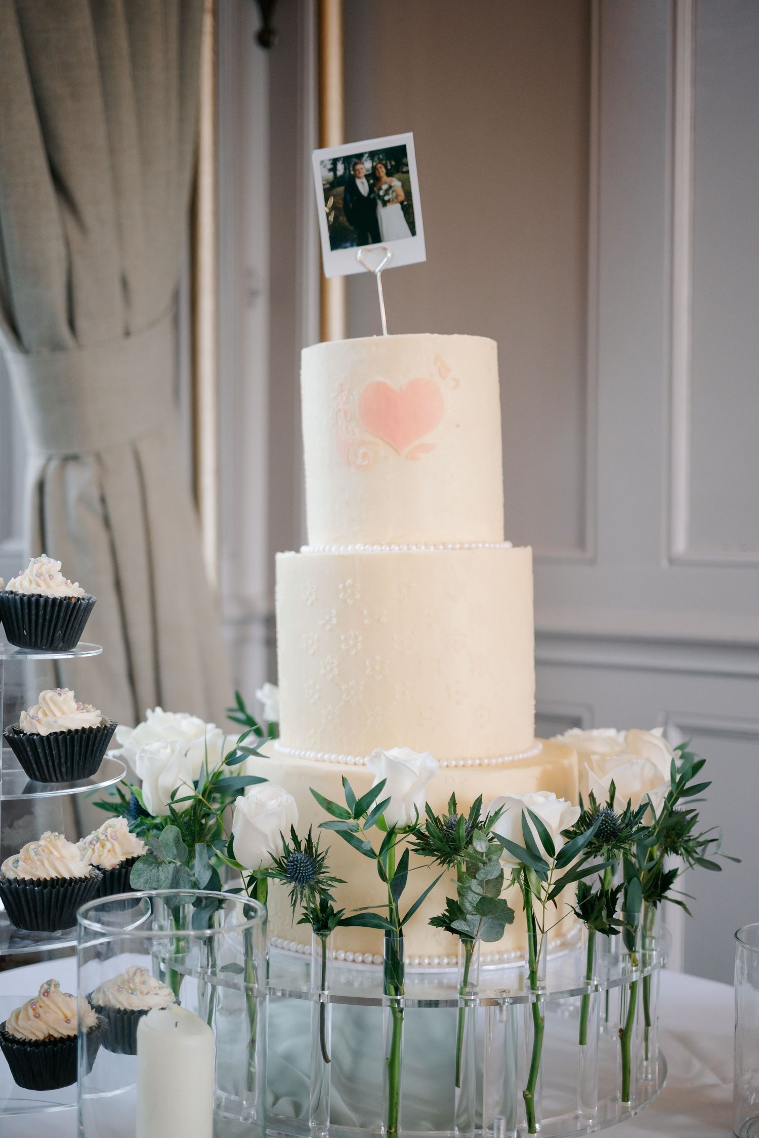 Elegant white wedding cake and cupcakes displayed with greenery and glass vases.