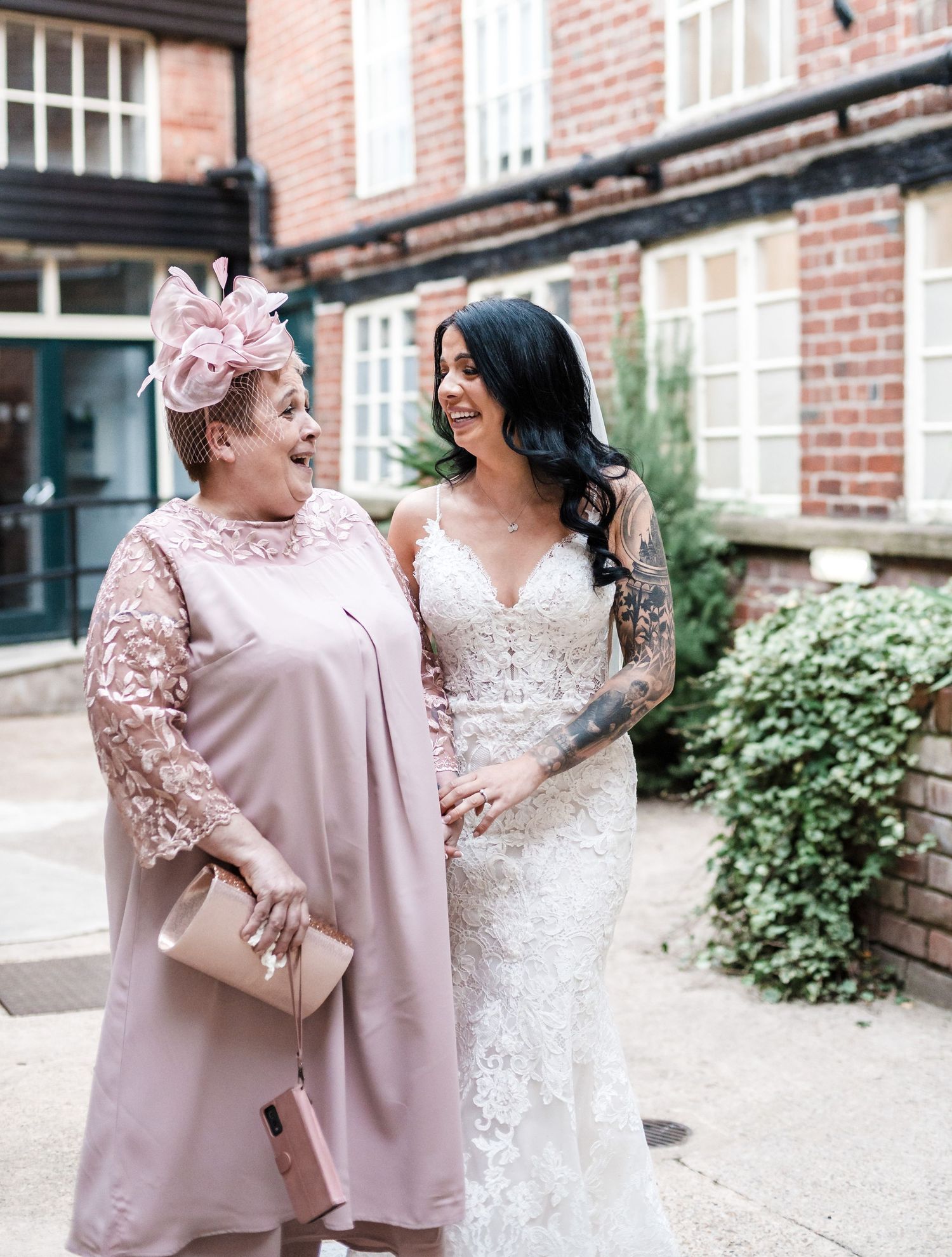 Two wedding guests share a joyful moment outside a brick building, one wearing a blush pink dress with fascinator.
