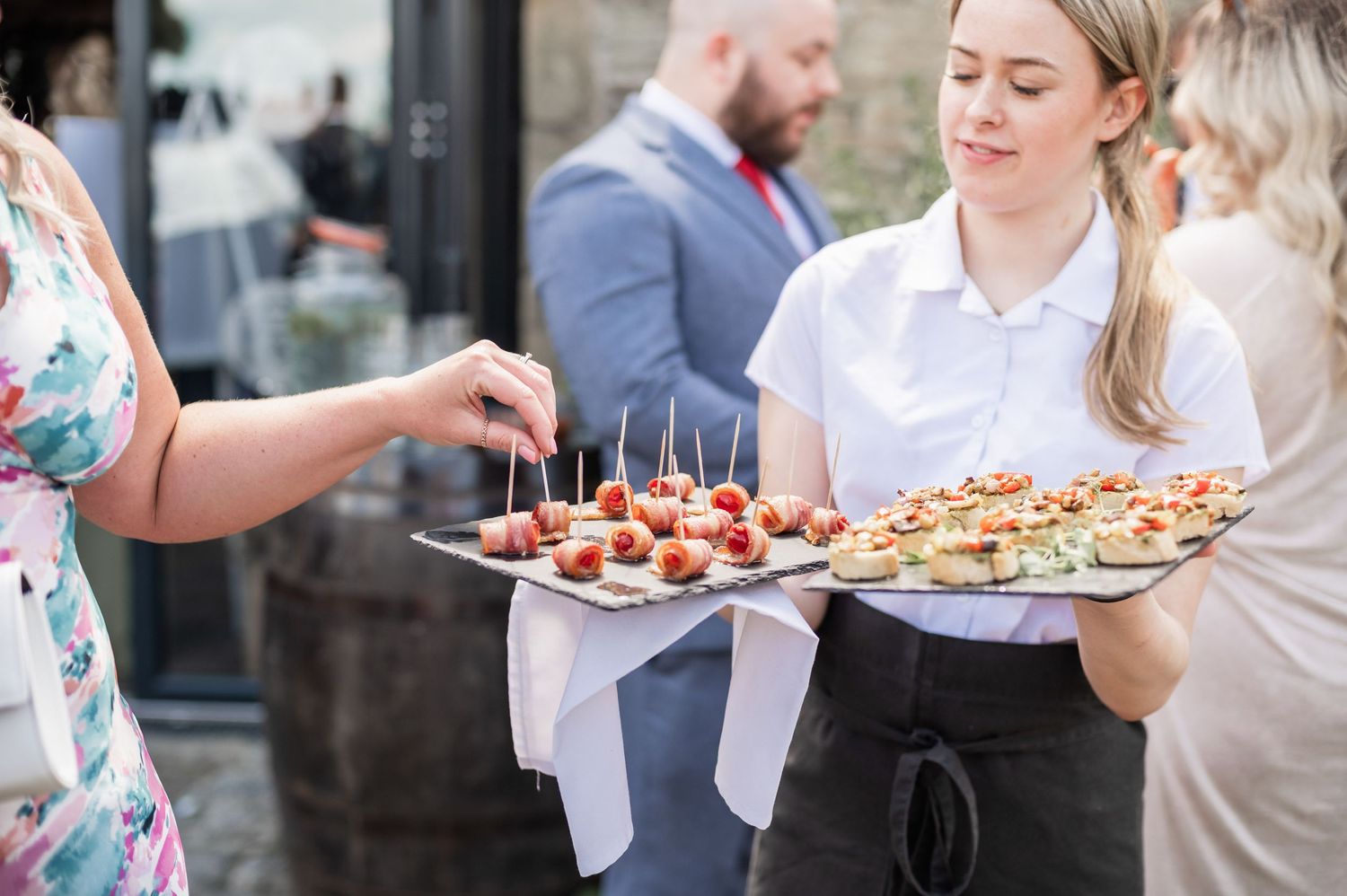 A server in professional attire presents appetizer platters at an outdoor event.
