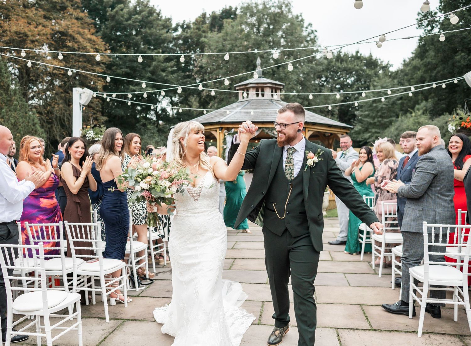 Newlyweds celebrate while walking down aisle under string lights as guests cheer.