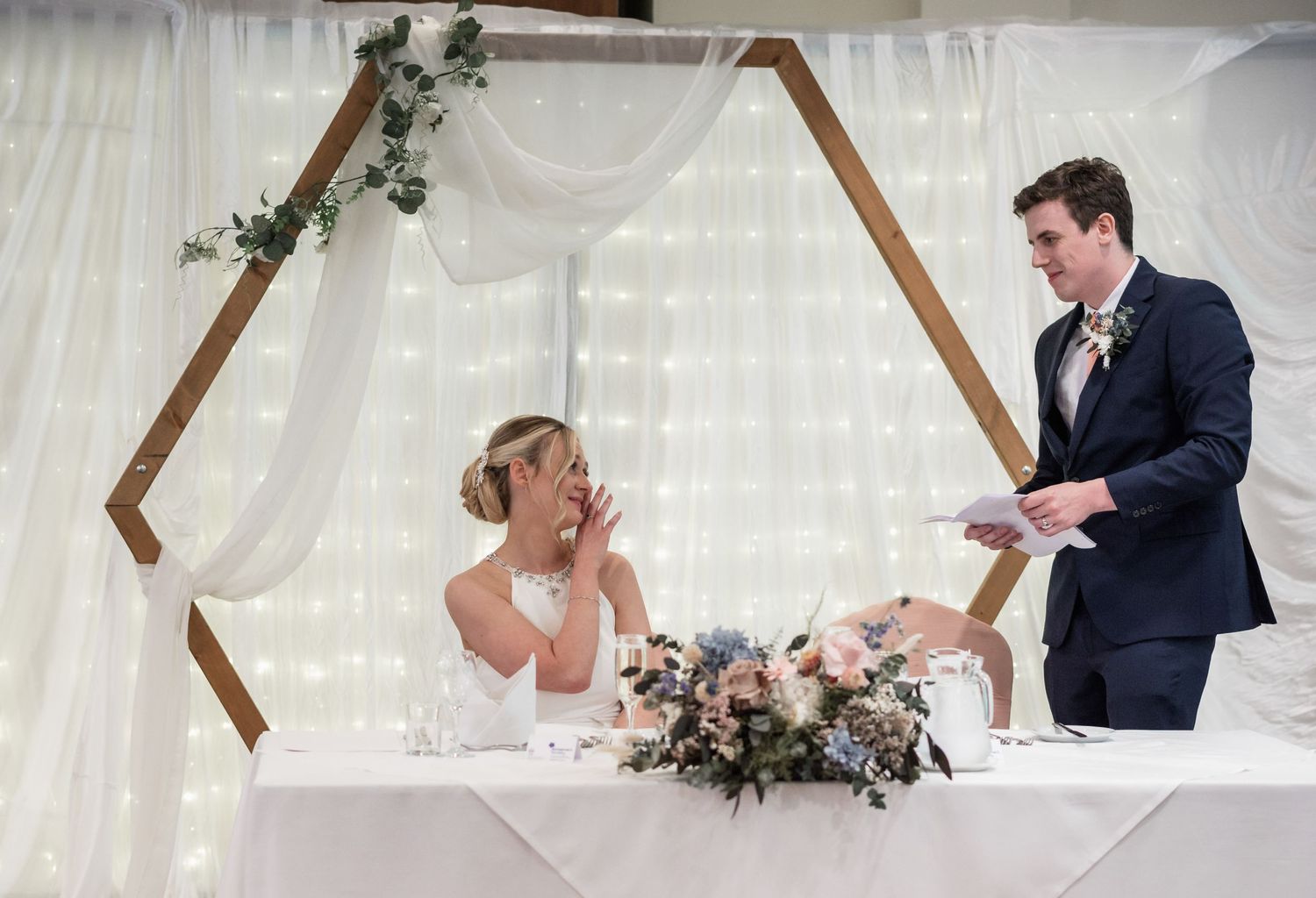 A candid moment during wedding speeches in a banquet hall with decorative white draping and wooden arch.