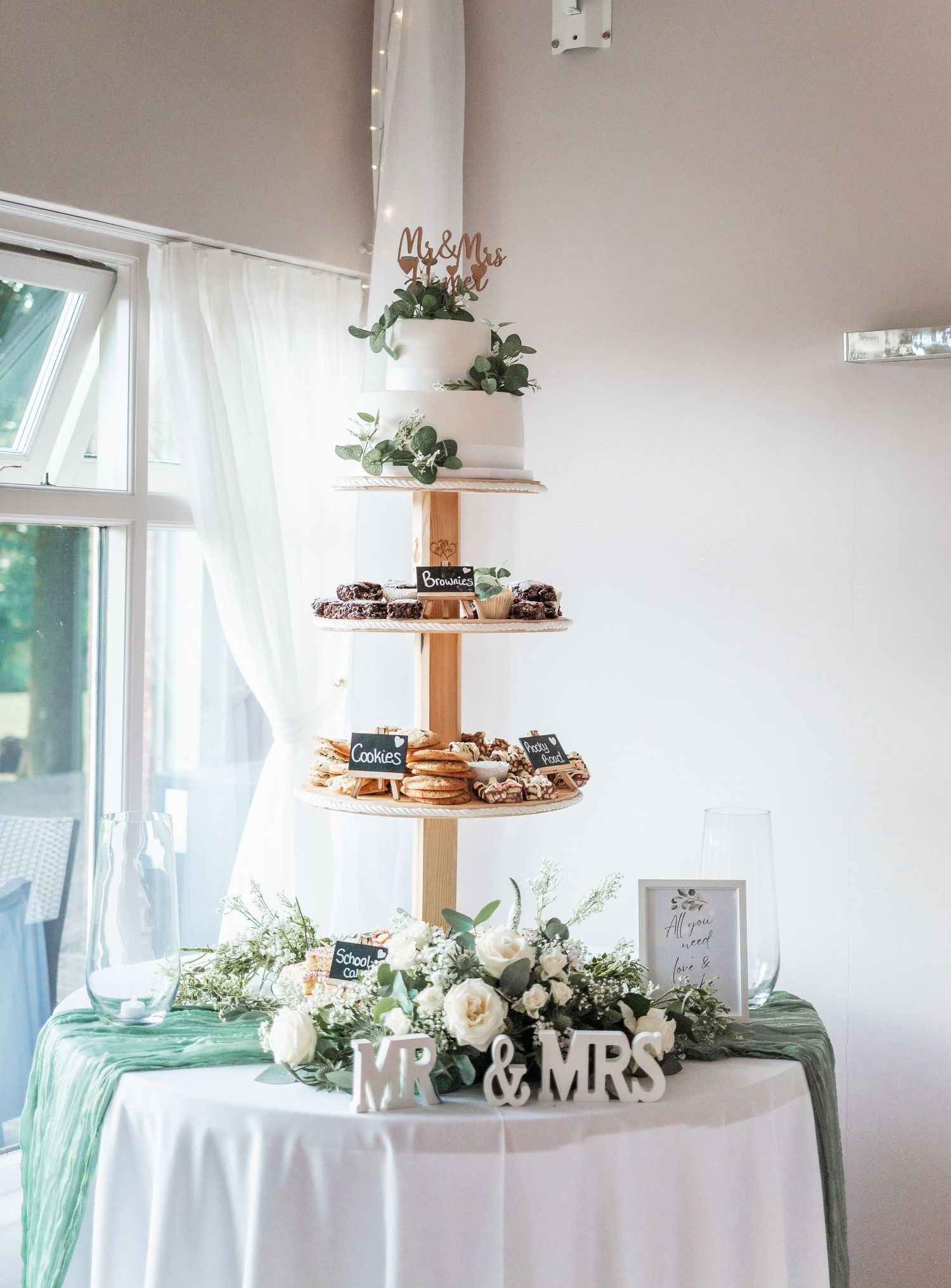 Elegant tiered dessert display with white cake and greenery on draped table with Mr & Mrs sign.