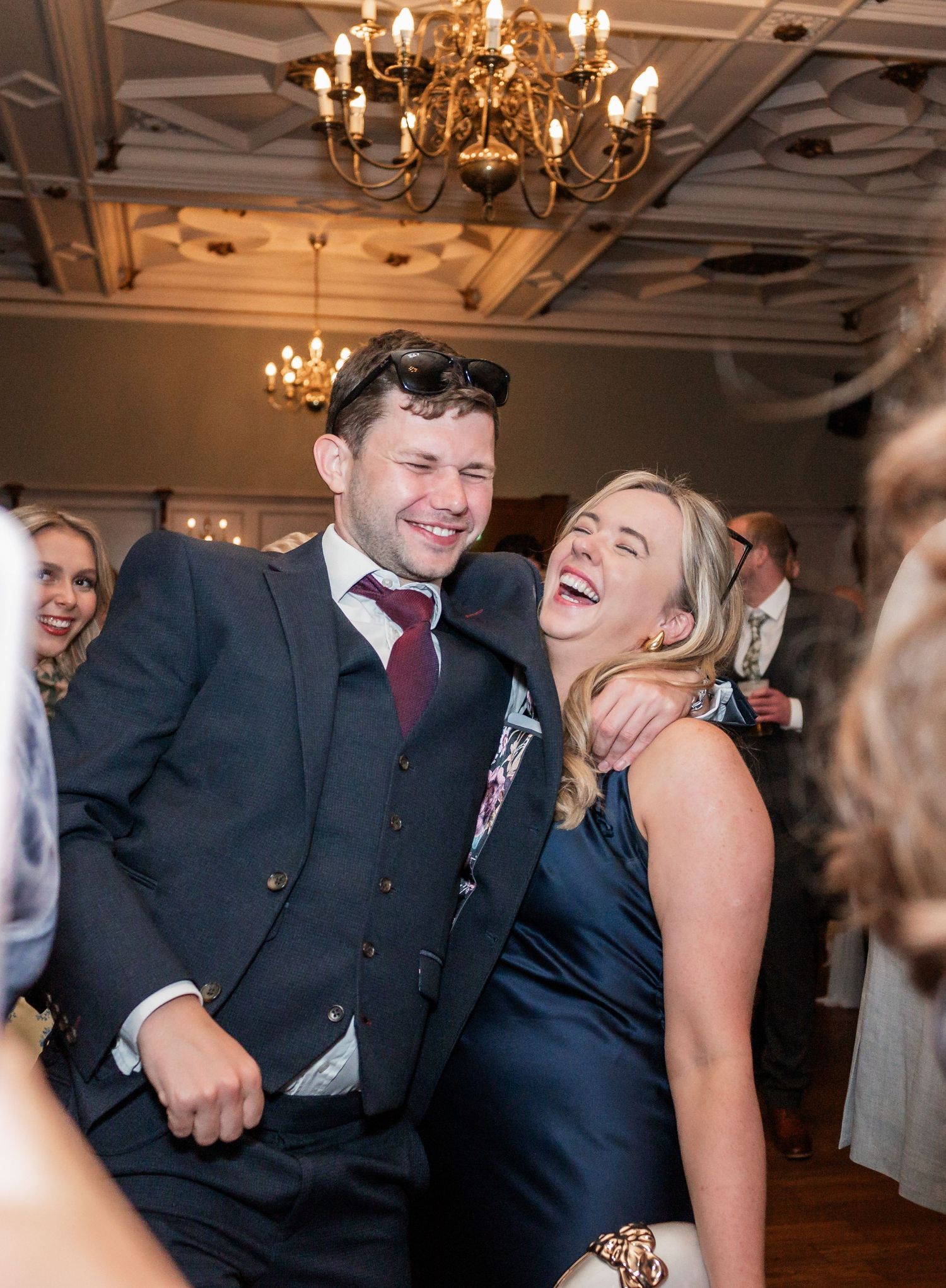 Wedding guests dance and celebrate on reception dance floor under ornate chandeliers.