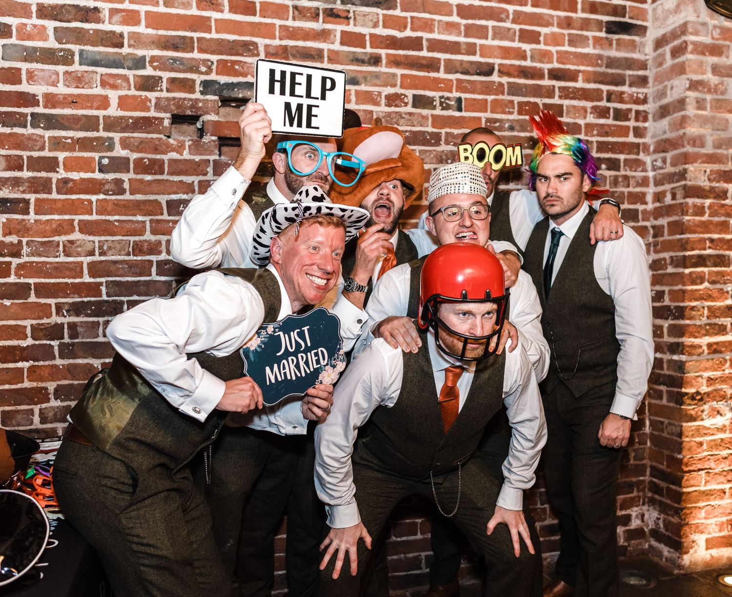 Wedding party poses with props and silly expressions against brick wall.