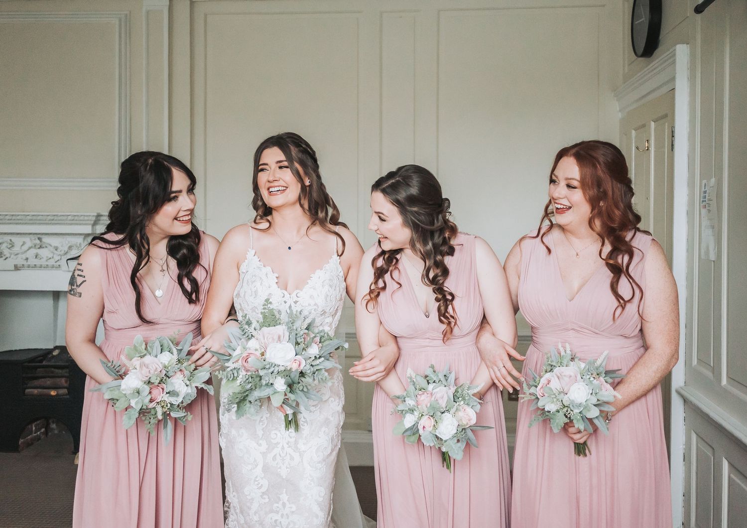 A wedding party poses with matching pink dresses and pastel bouquets in an elegant white room.