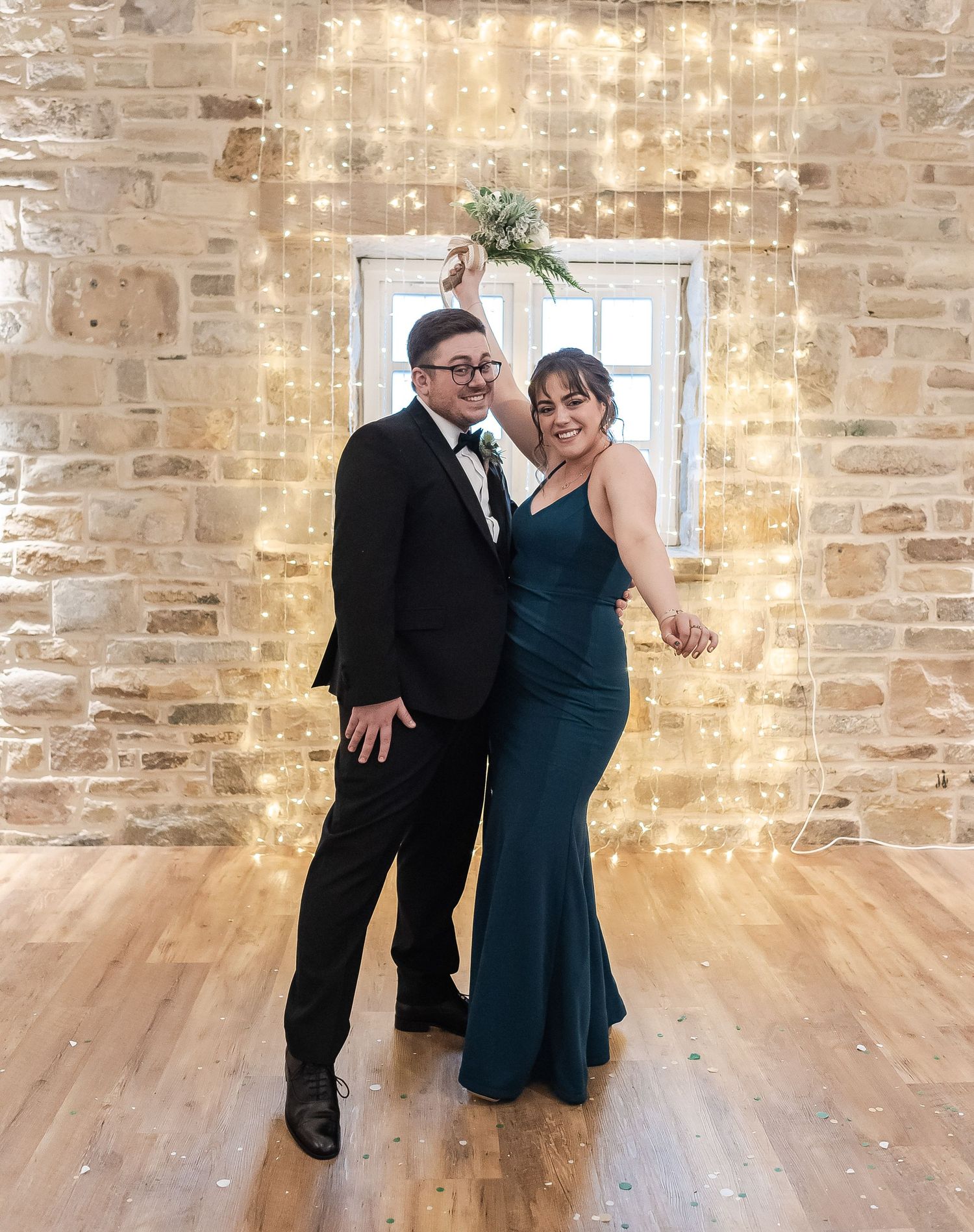 Formal couple posing against stone wall with twinkling fairy lights backdrop.