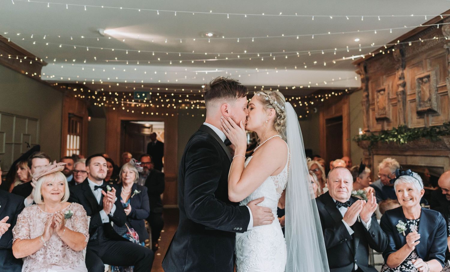 Newlyweds share first kiss while guests applaud under twinkling lights at indoor reception.