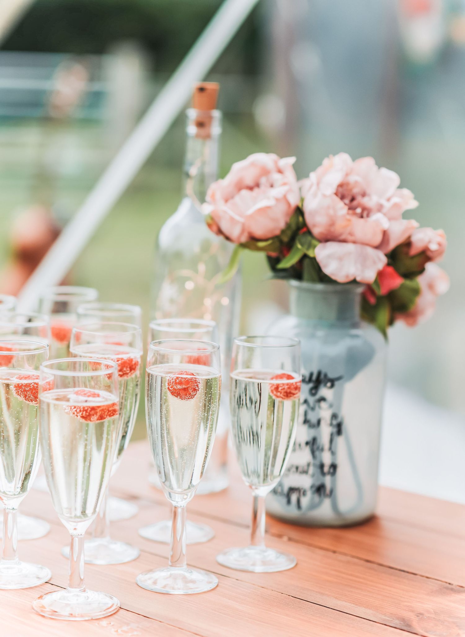 Champagne flutes and pink peonies arranged on wooden table at outdoor event.