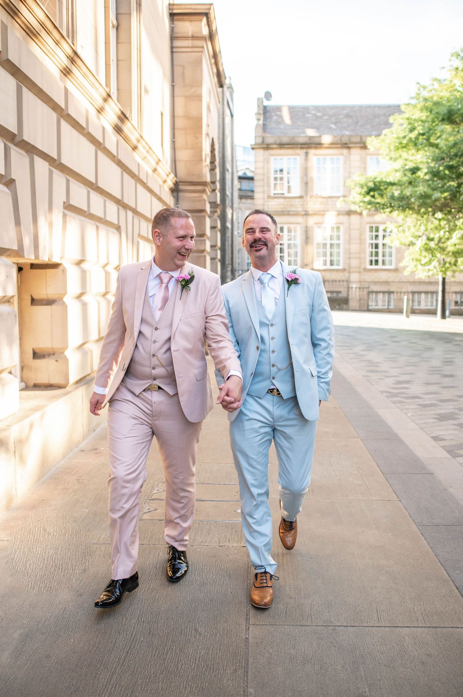 Happy couple in pink and blue suits walking hand in hand along a stone building.